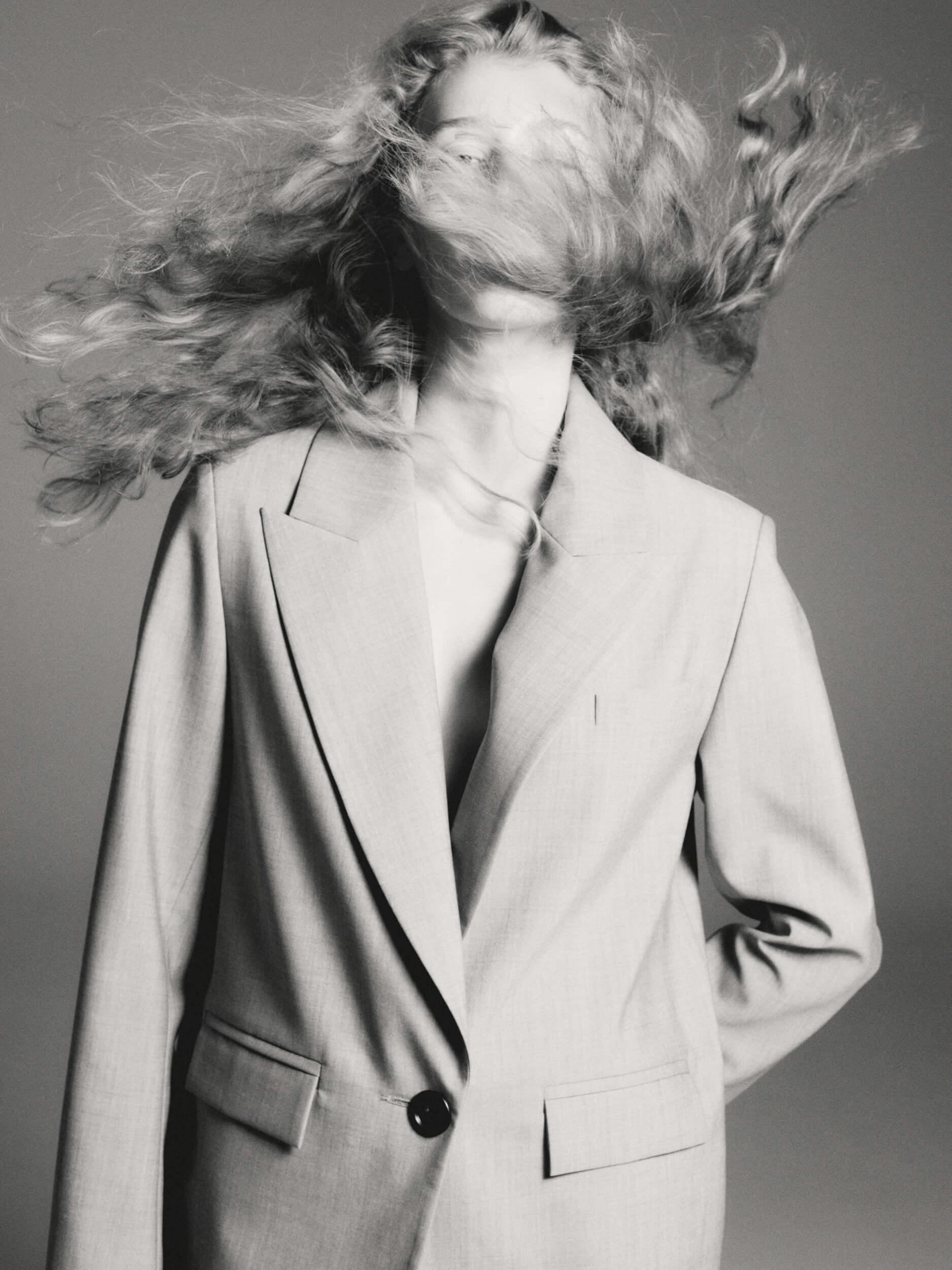 A person wearing a light-colored blazer stands with their head tilted slightly back as their long, wavy hair sweeps across their face. The image is in black and white with a plain background.