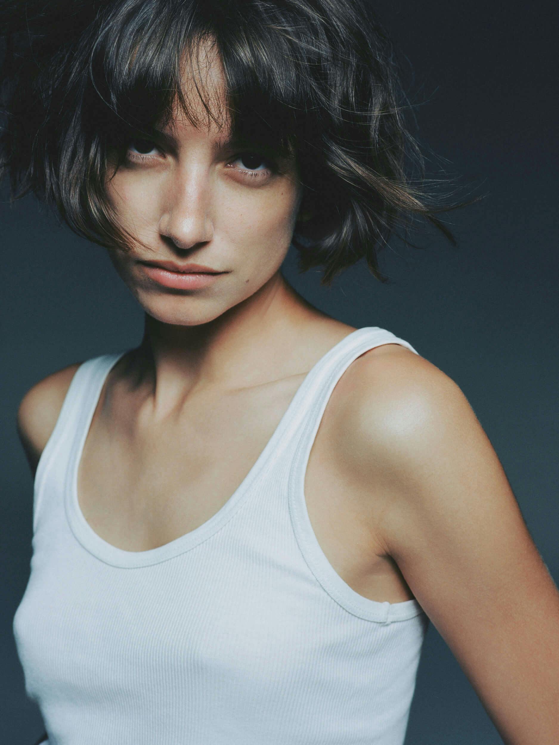 A person with short, tousled brown hair and bangs, wearing a white tank top, looks toward the camera against a dark background. Their expression is neutral.