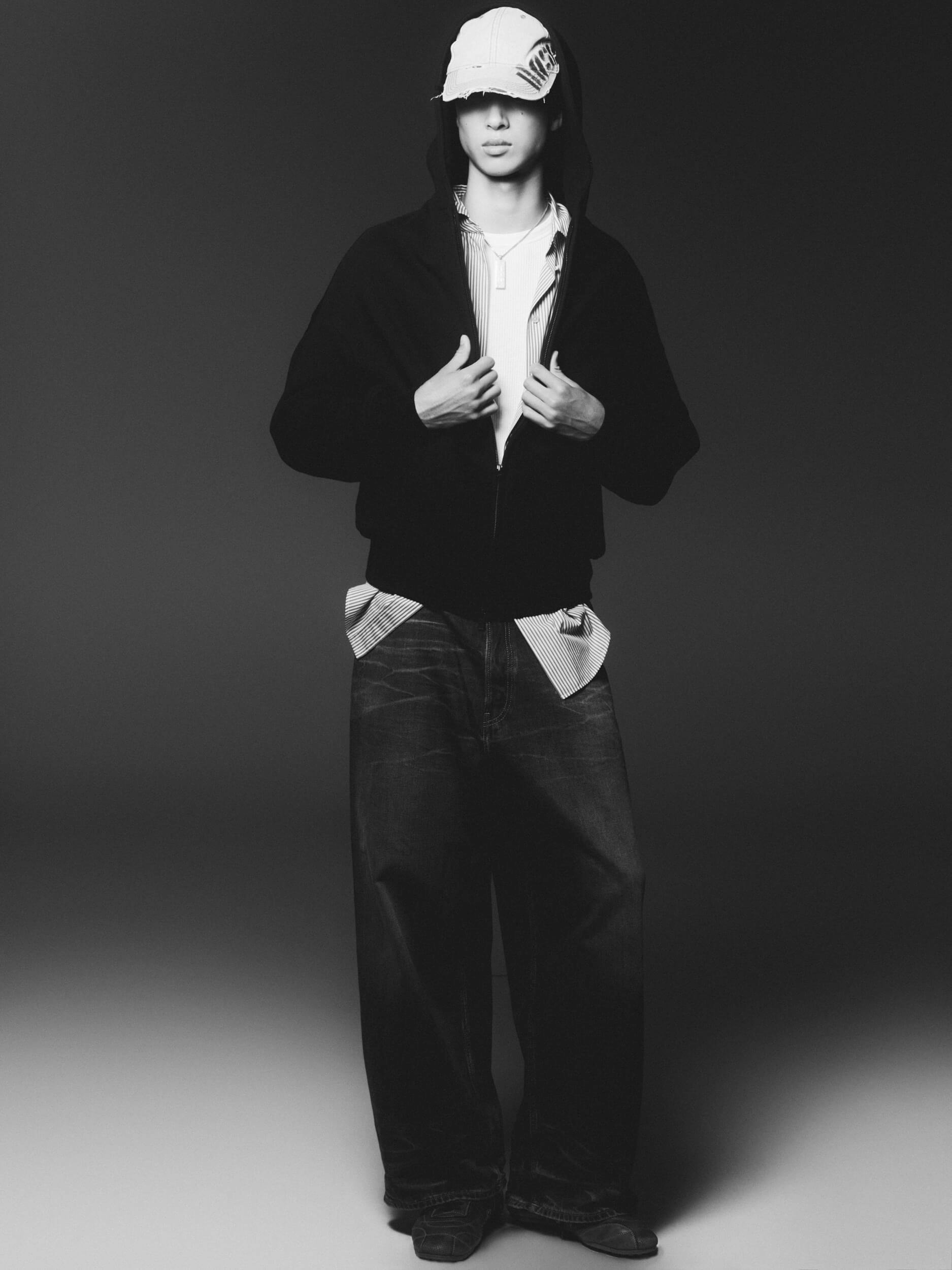 A person stands against a plain background wearing a hooded jacket, a white shirt, a white cap, and loose-fitting dark jeans. The image is in black and white. The person faces forward with hands on the jacket.