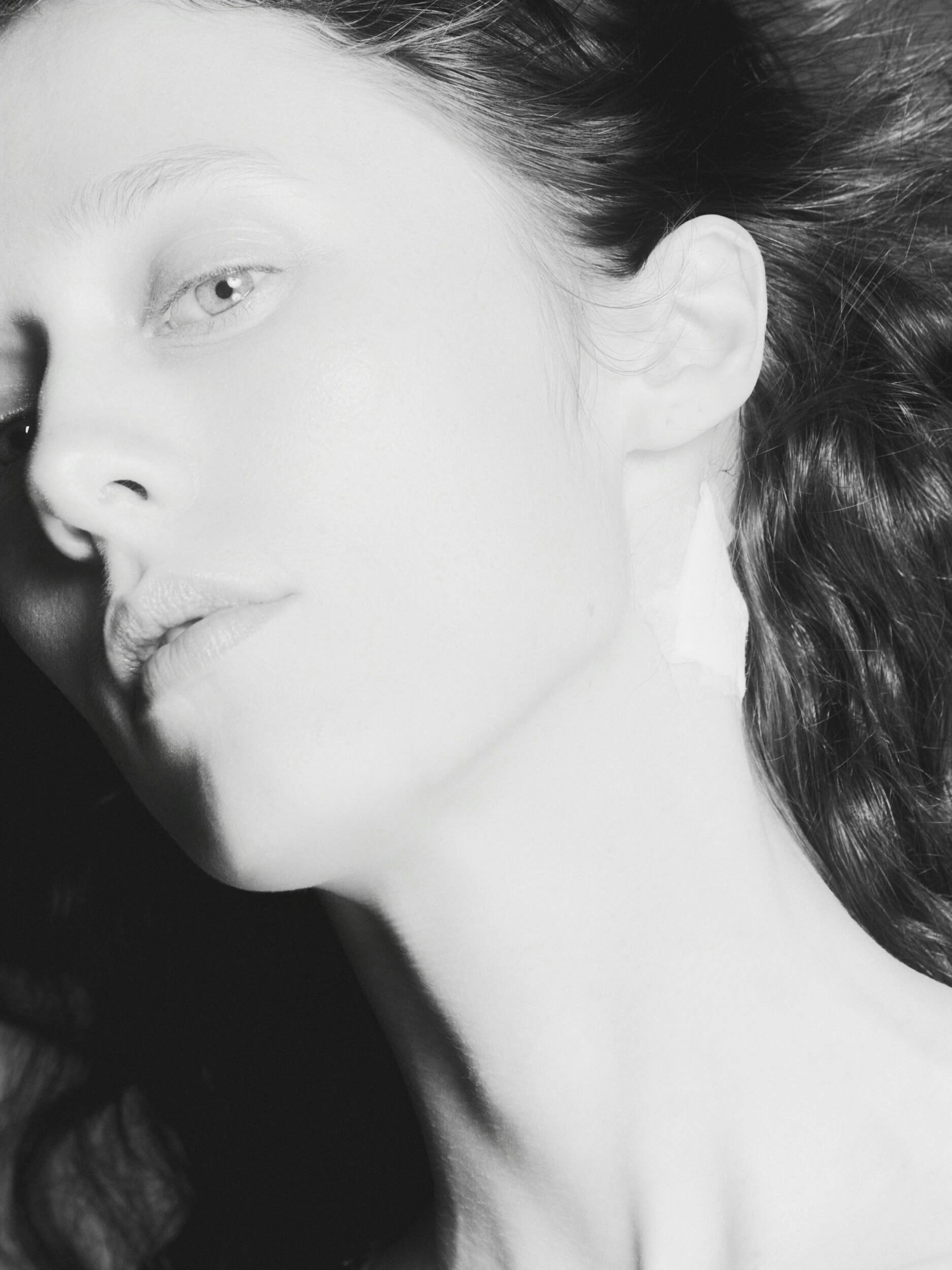 A close-up black and white photo of a person's face in profile, showing pale skin, light eyes, and slightly parted lips. The person has long, wavy hair and a small piece of white tape behind their ear.