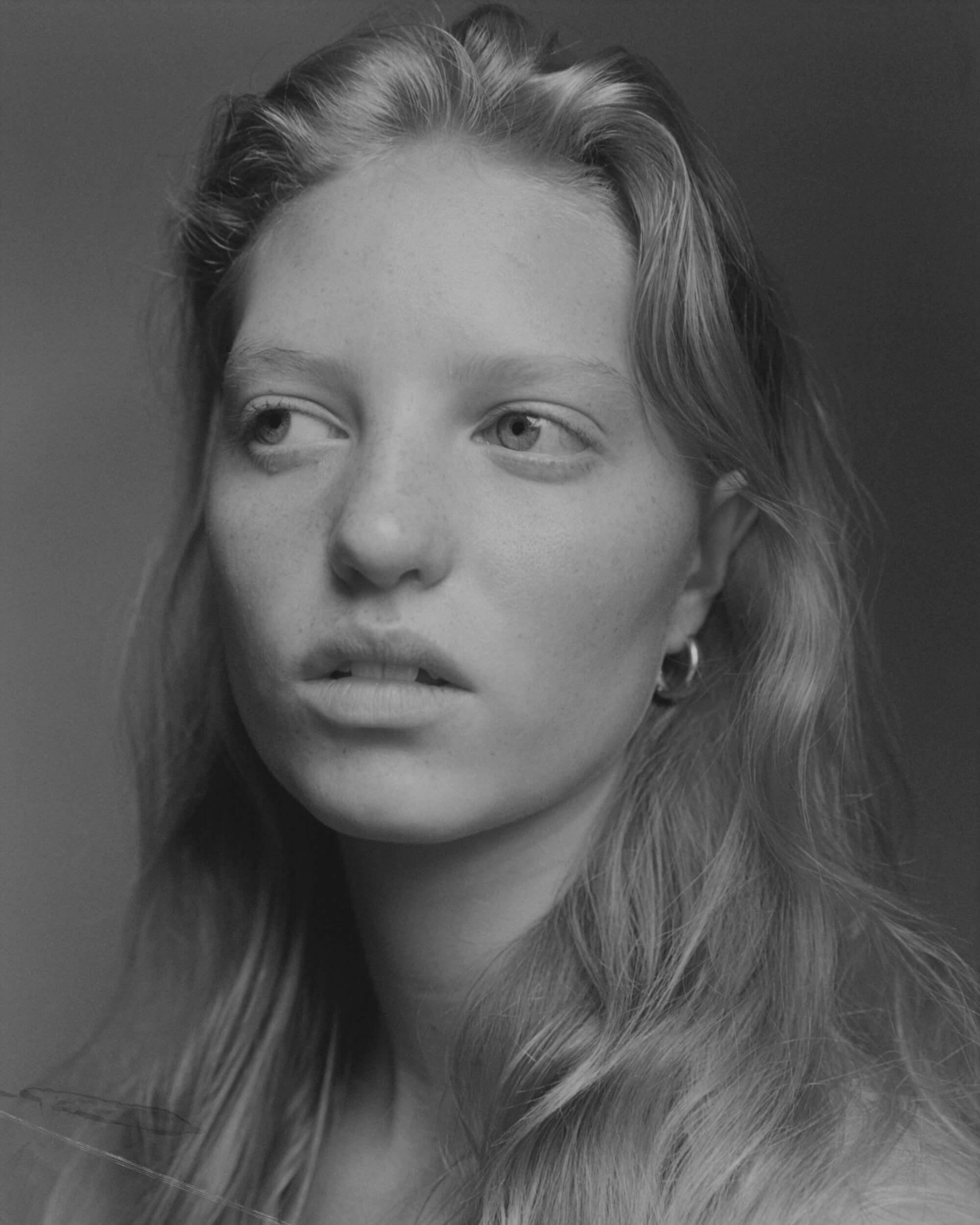 A black and white portrait of a young person with long, wavy hair, looking slightly to the left. They have light skin, minimal makeup, and are wearing hoop earrings. The background is plain and softly lit.