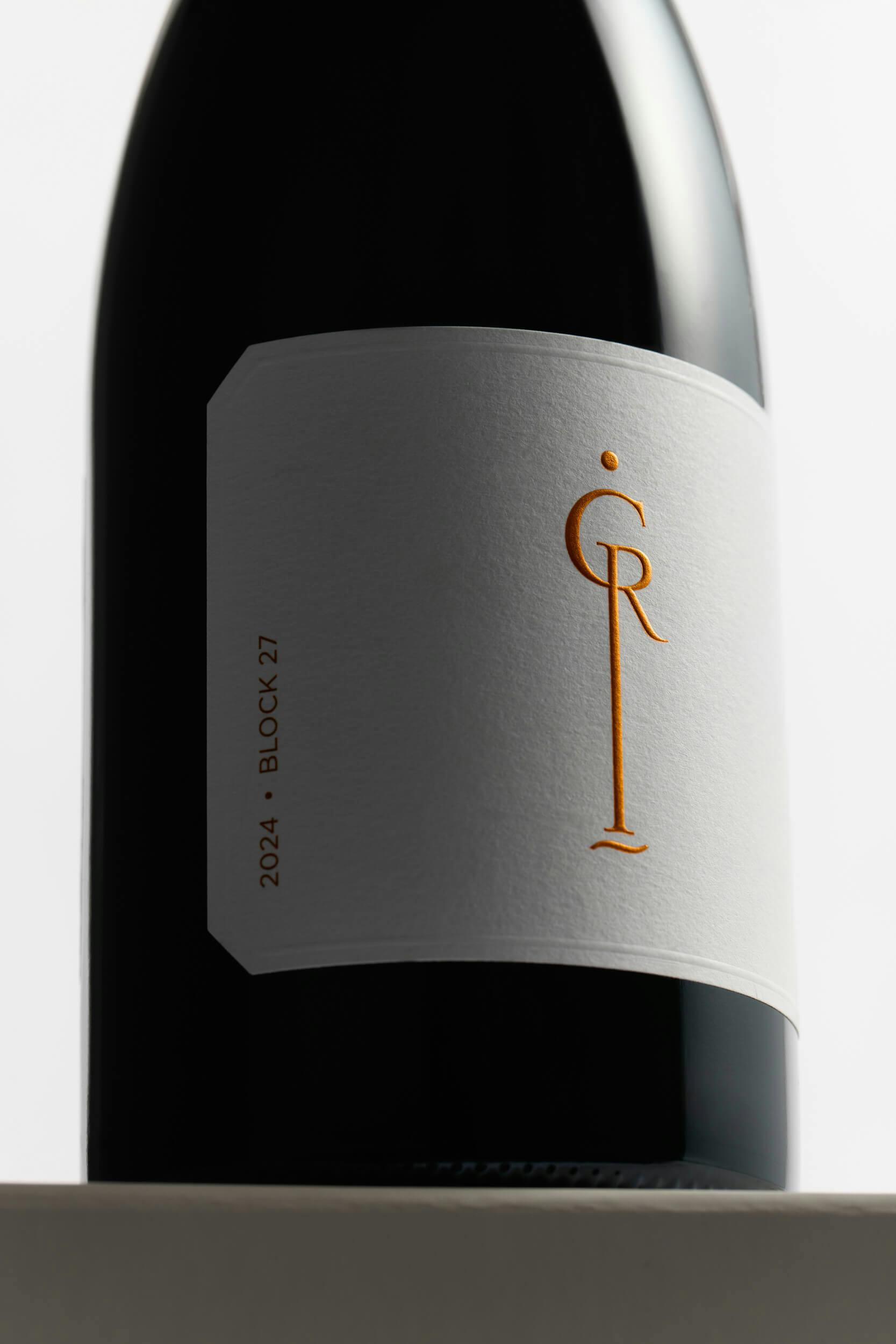 A close-up of a dark wine bottle with a minimal white label featuring gold lettering. The label shows 