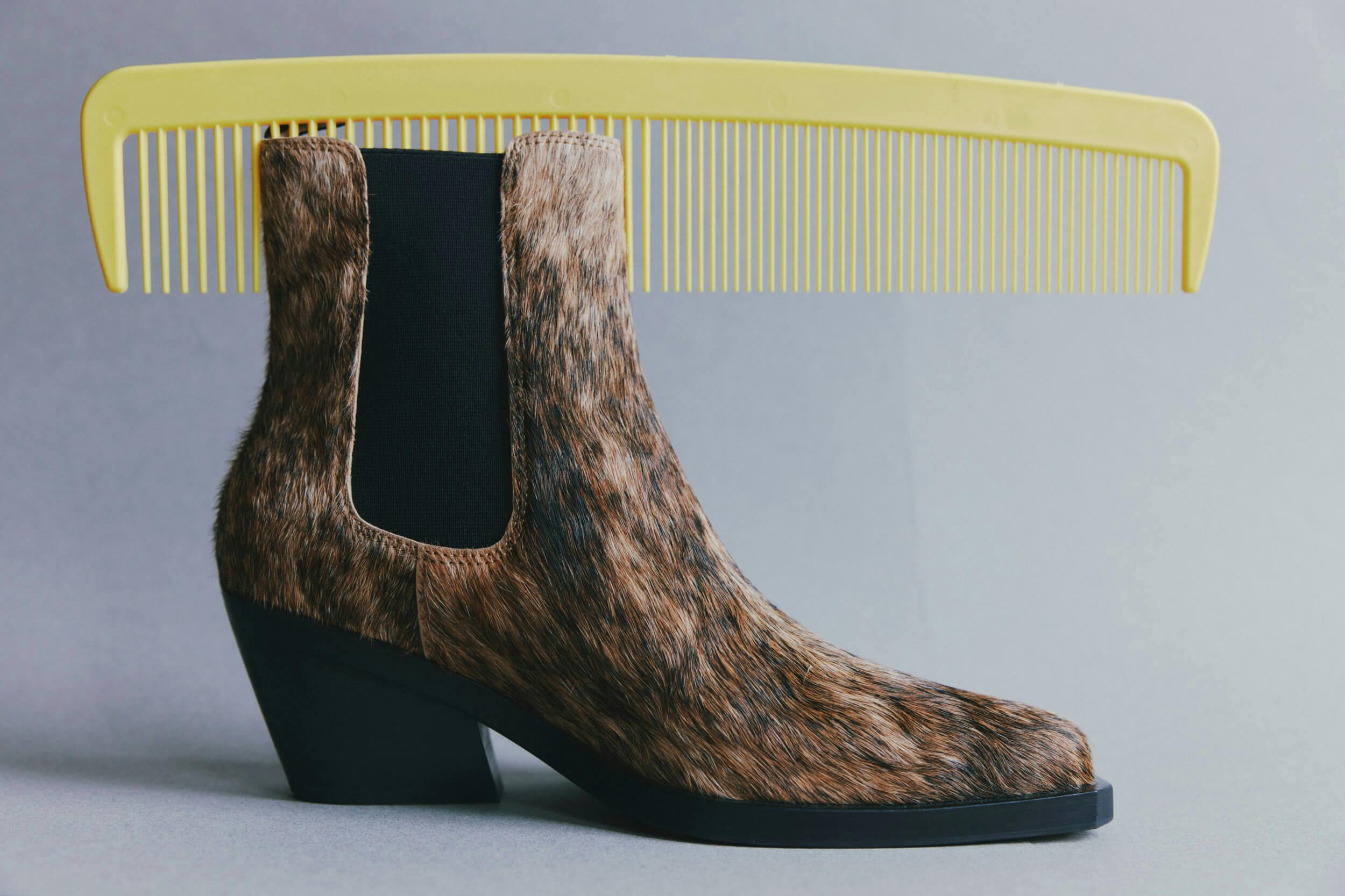 A brown, faux fur ankle boot with a black elastic side panel is positioned in front of a large yellow comb, both set against a plain gray background.