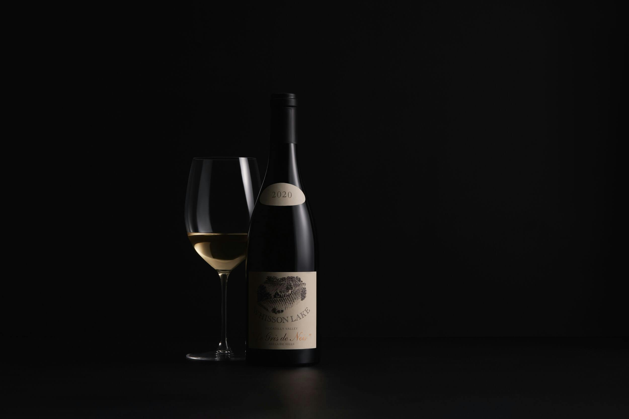 A bottle of white wine with a detailed label stands next to a filled wine glass against a plain, dark background. The lighting highlights the glass and the front of the bottle.