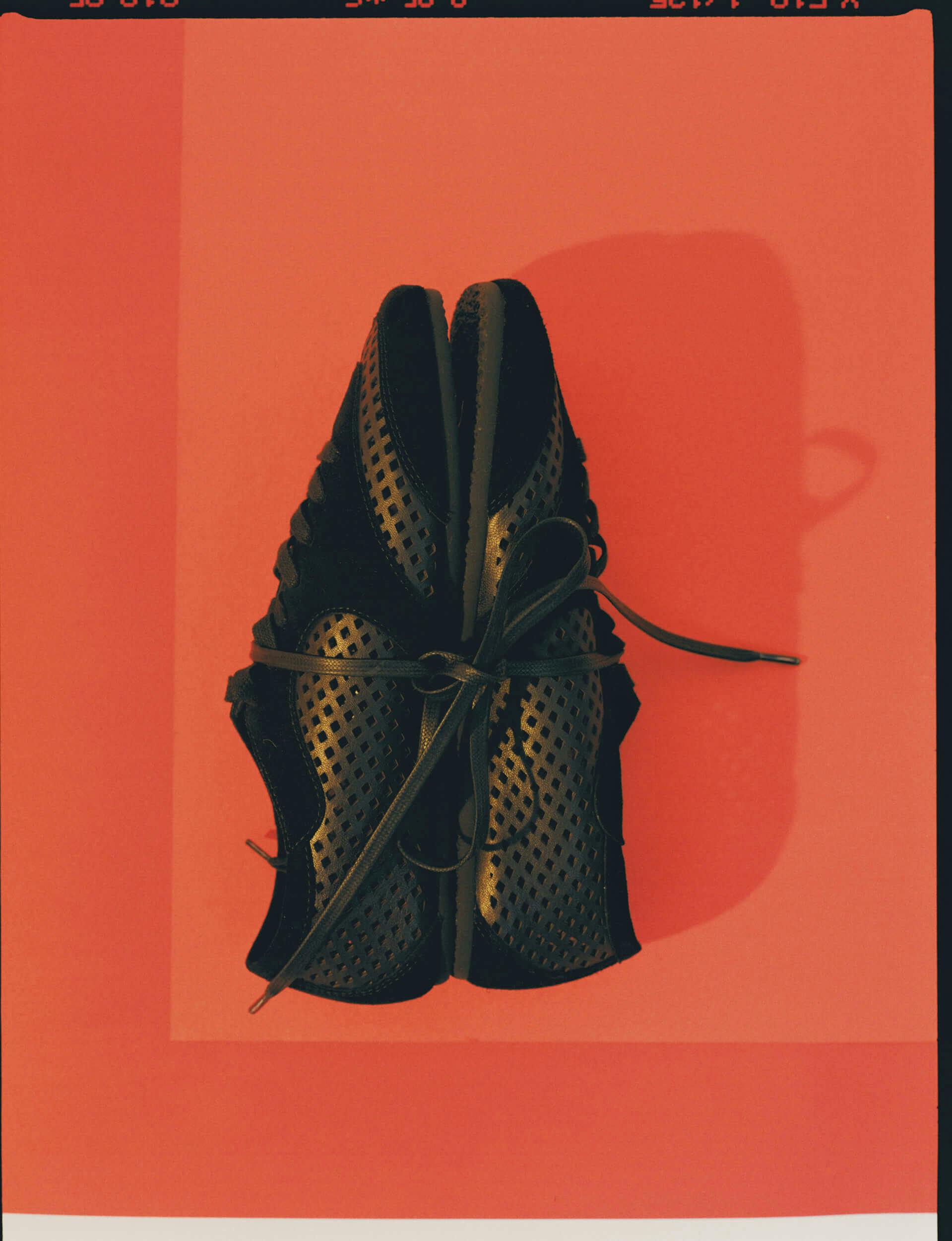 A pair of black, perforated athletic shoes are placed side by side with their laces tied together, viewed from above on a red background with a lighter red rectangular border.