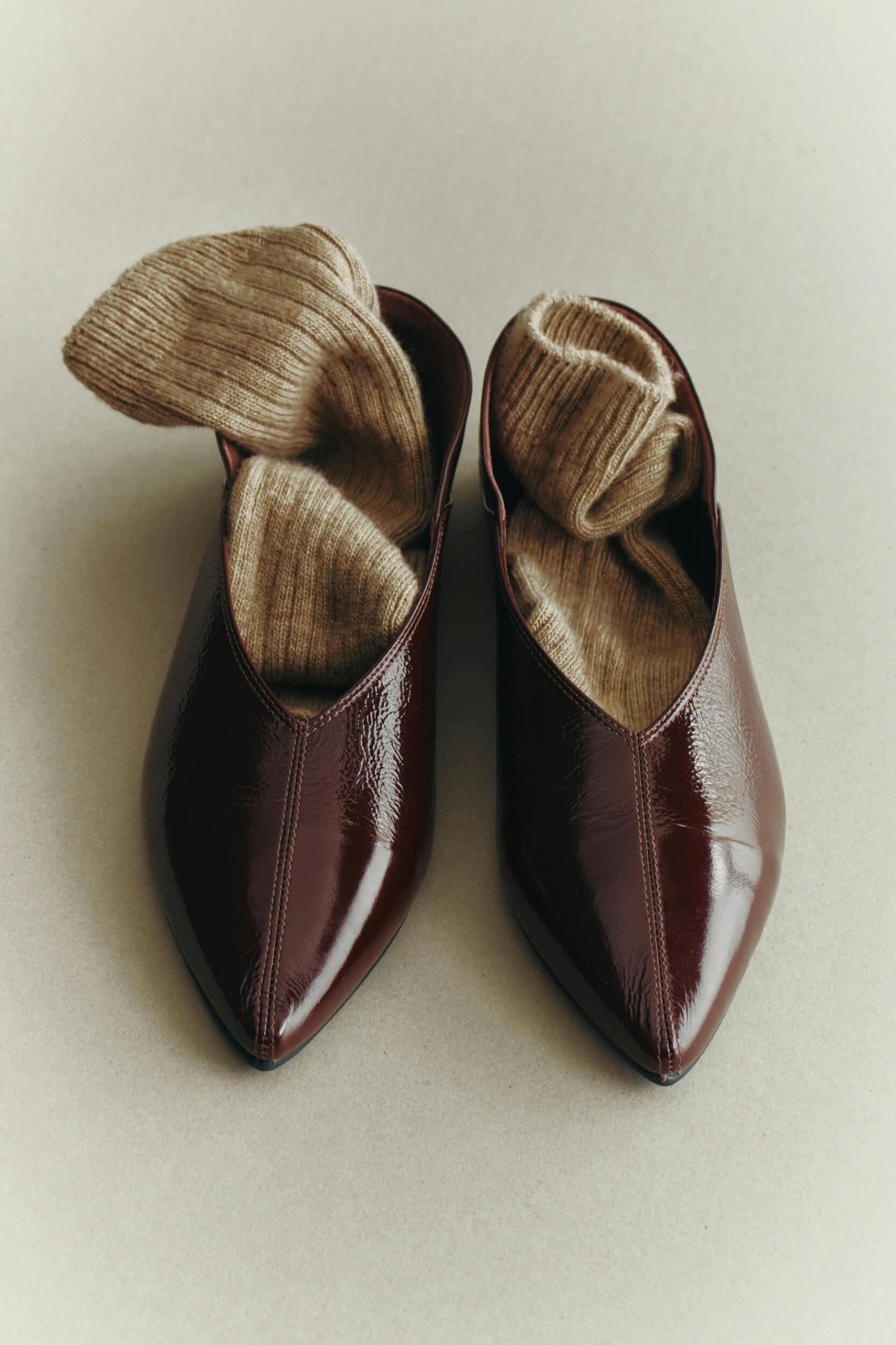 A pair of pointed, dark brown leather shoes with a glossy finish, each containing a rolled beige knit sock, placed on a plain light-colored surface.