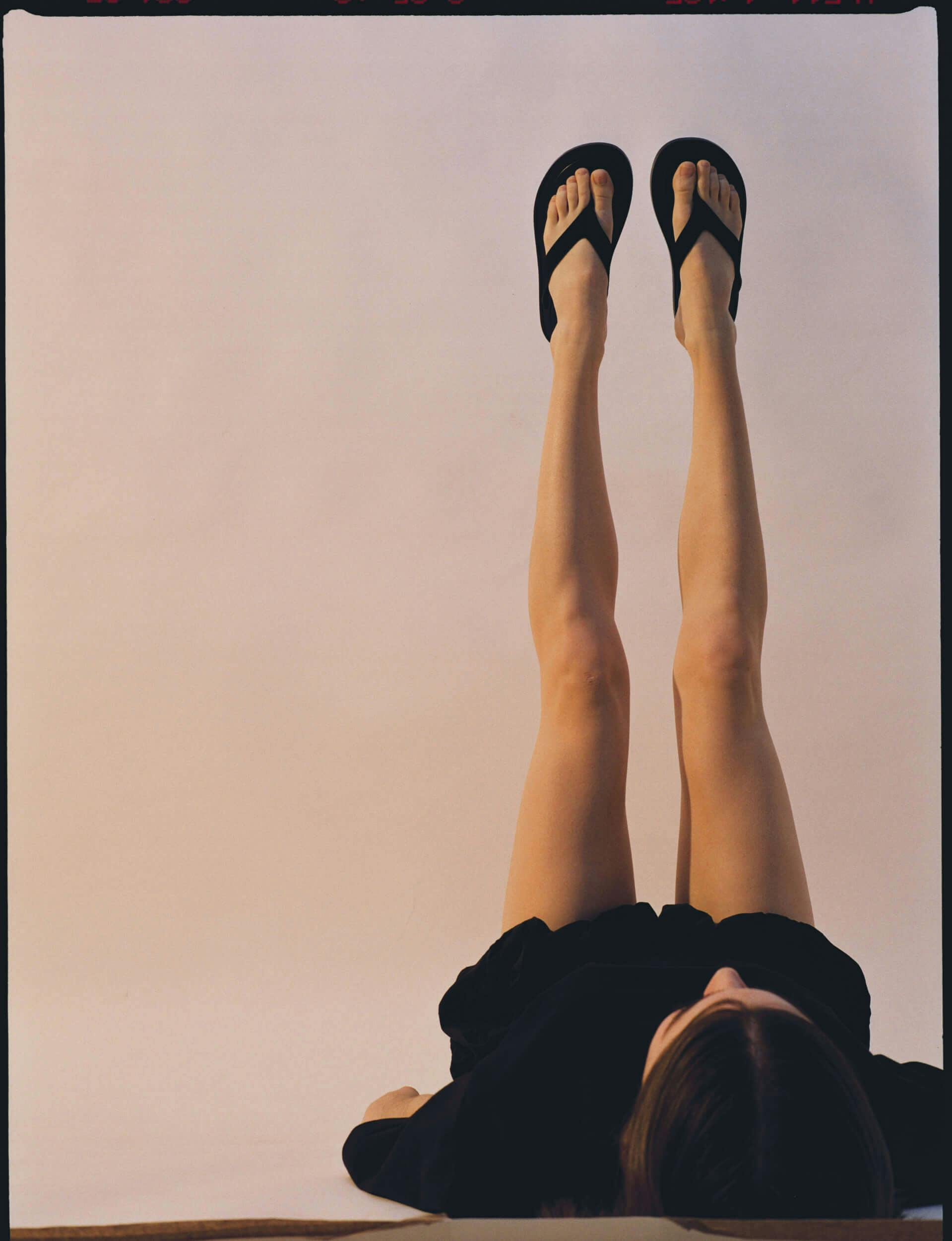 A person wearing a black outfit lies on their back with legs extended upward, showing black flip flops against a plain, light-colored background.