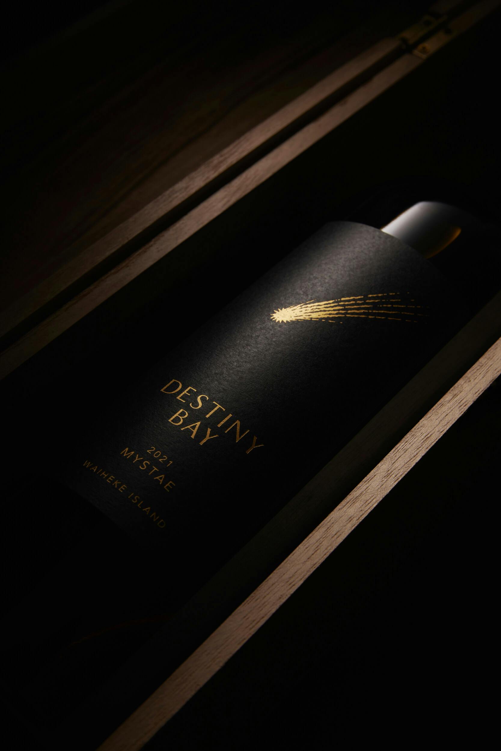 A bottle of Destiny Bay wine in a wooden box, featuring a black label with gold text and a comet illustration. The label reads “Destiny Bay 2022 Mystae Waiheke Island.”.