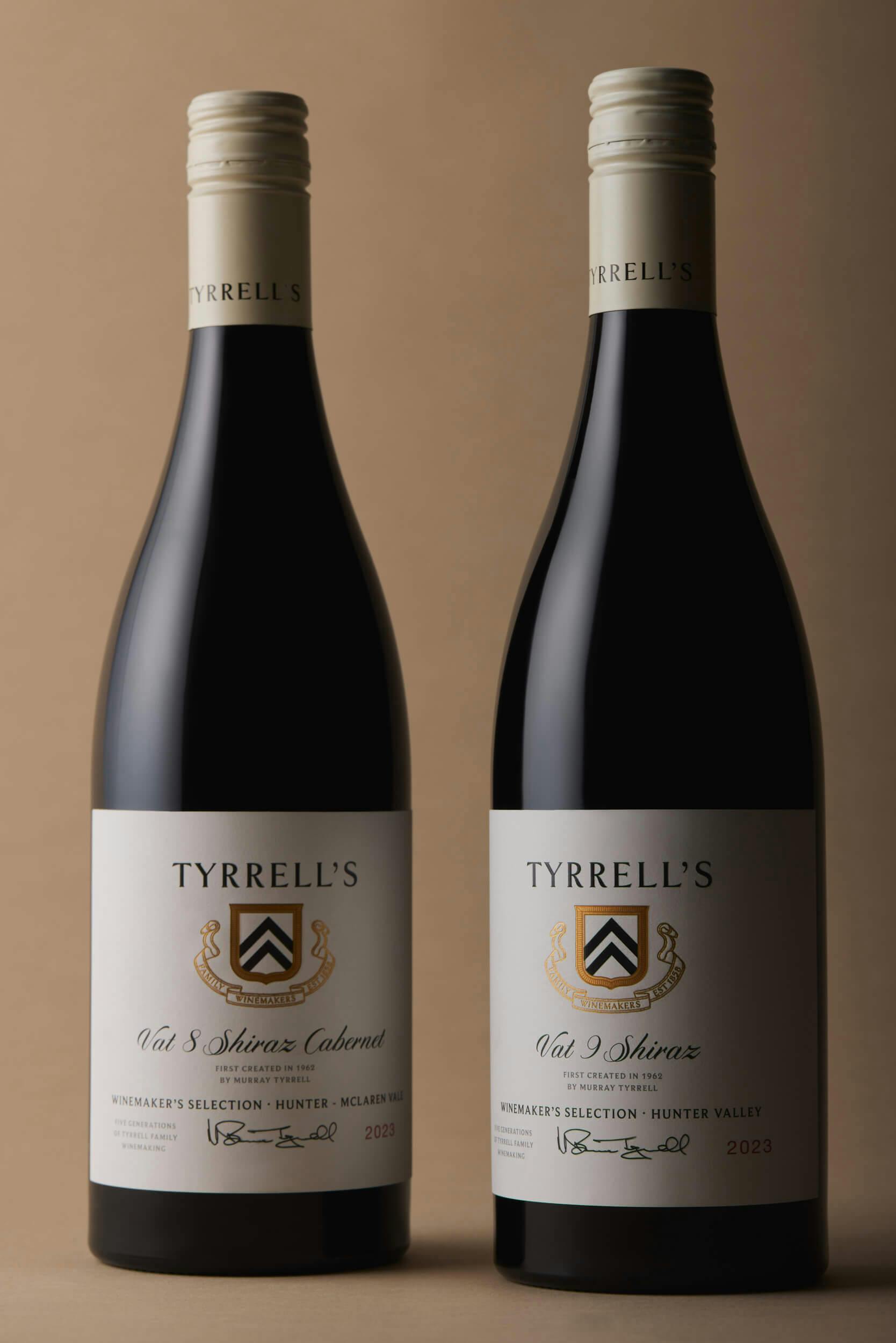 Two bottles of Tyrrell's red wine stand side by side against a tan background. Both have cream-colored labels, gold and black crest designs, and black caps. The wine types and years are printed on the labels.