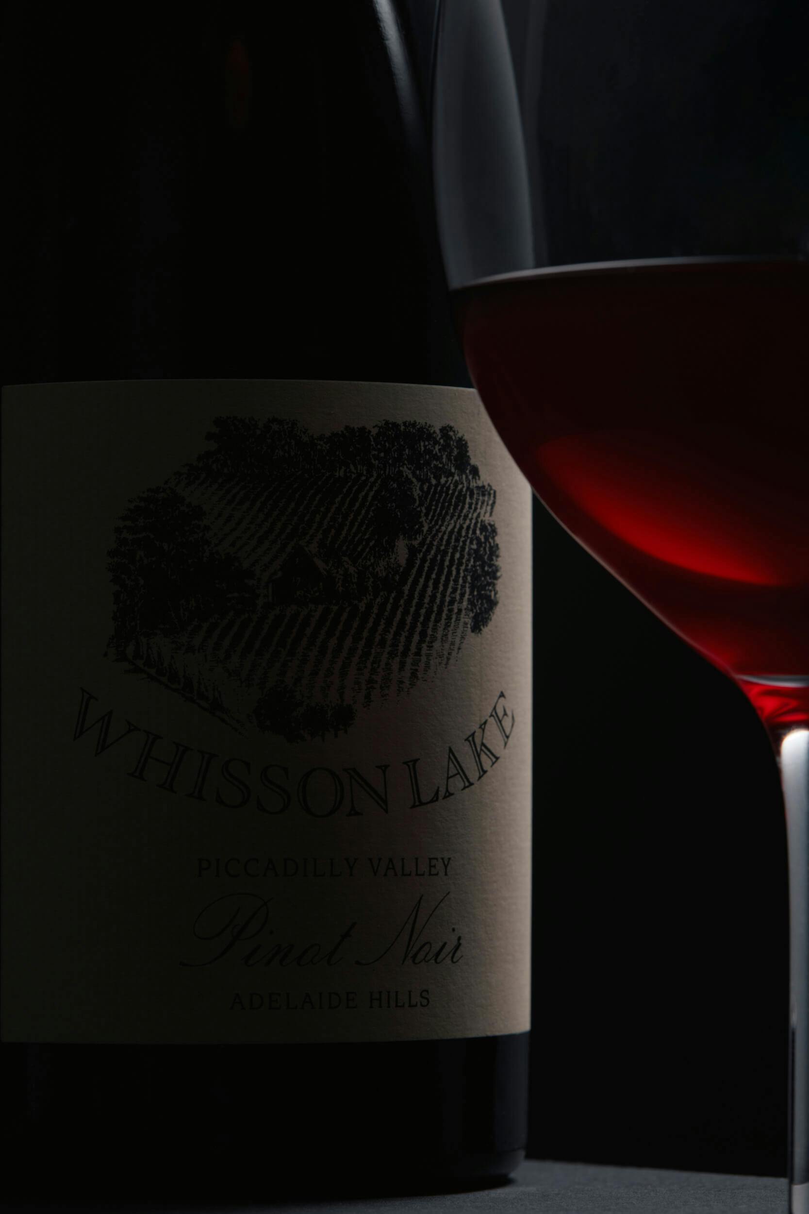 A bottle of Whisson Lake Pinot Noir wine next to a glass filled with red wine, both set against a dark background. The bottle label reads 
