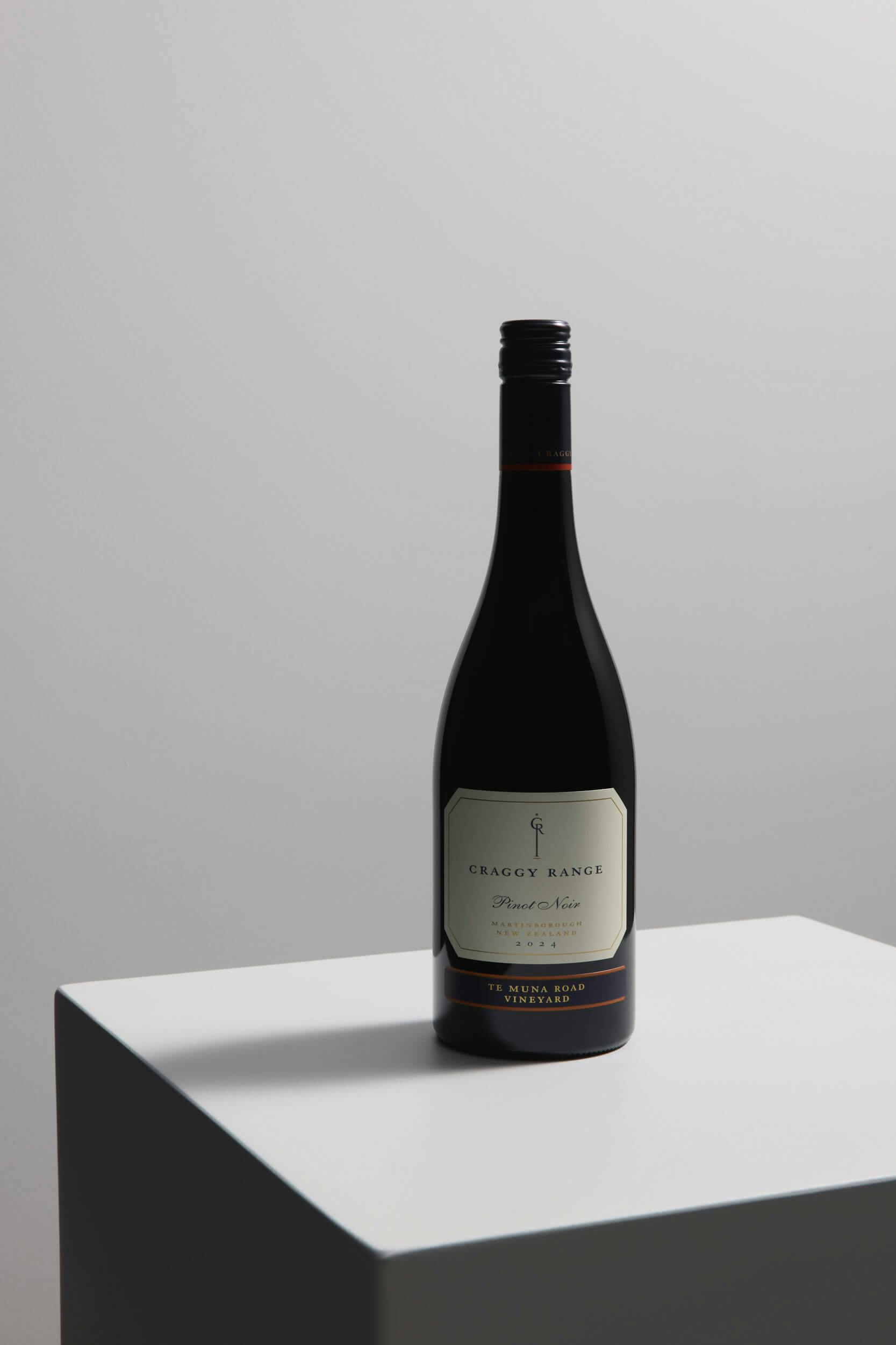 A bottle of Craggy Range Pinot Noir wine with a cream-colored label sits on a white, geometric pedestal against a plain, light gray background.