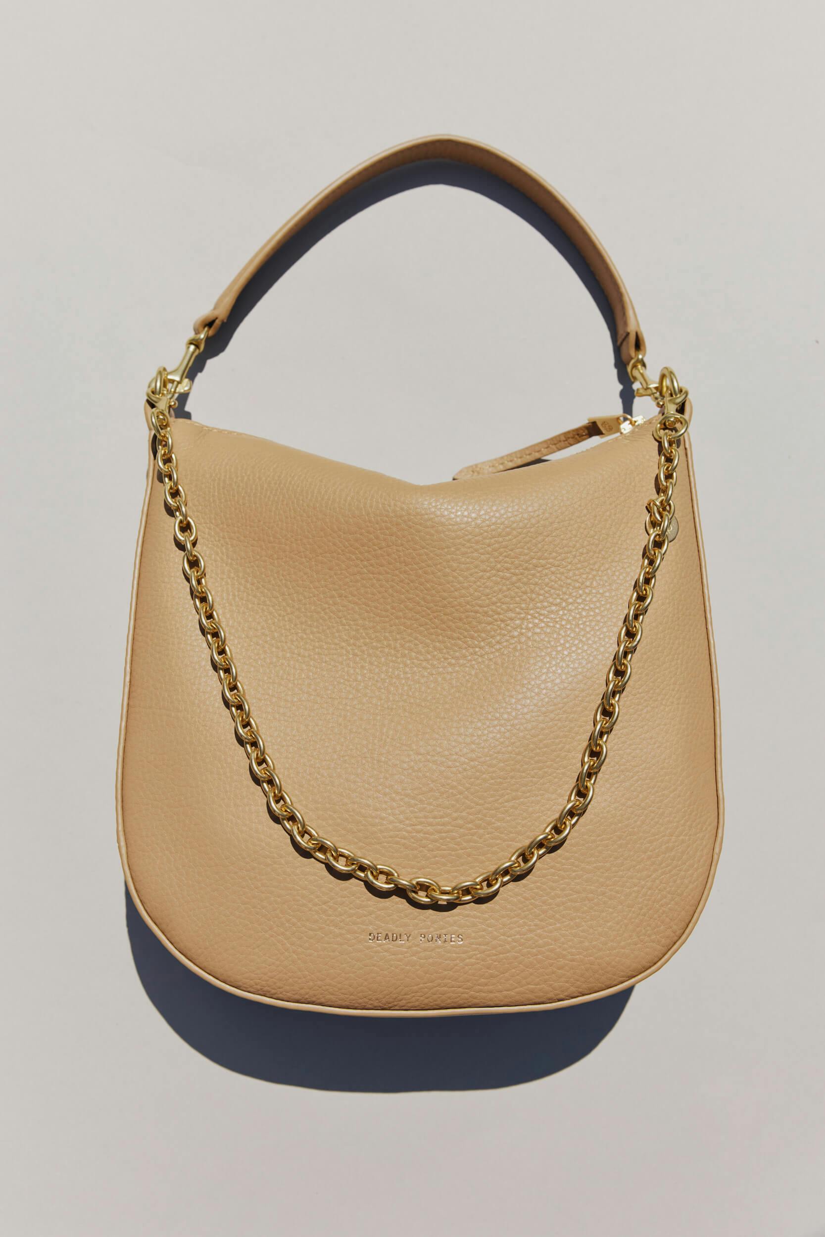 A tan leather handbag with a gold chain handle and a short strap is displayed against a light background. The bag has a pebbled texture and the brand name is embossed near the bottom.