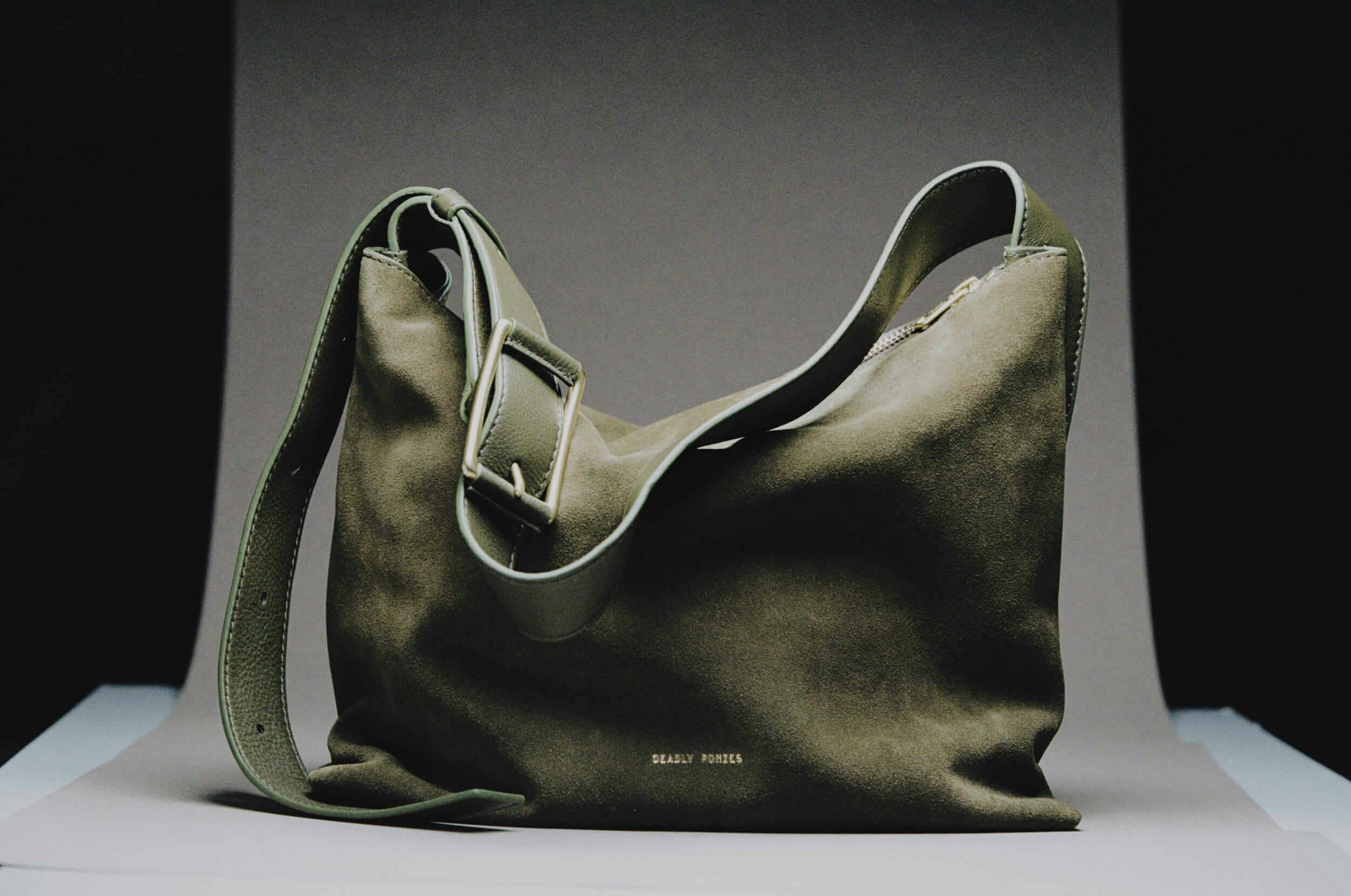 A large olive-green suede shoulder bag with a thick adjustable strap and subtle gold lettering reading 