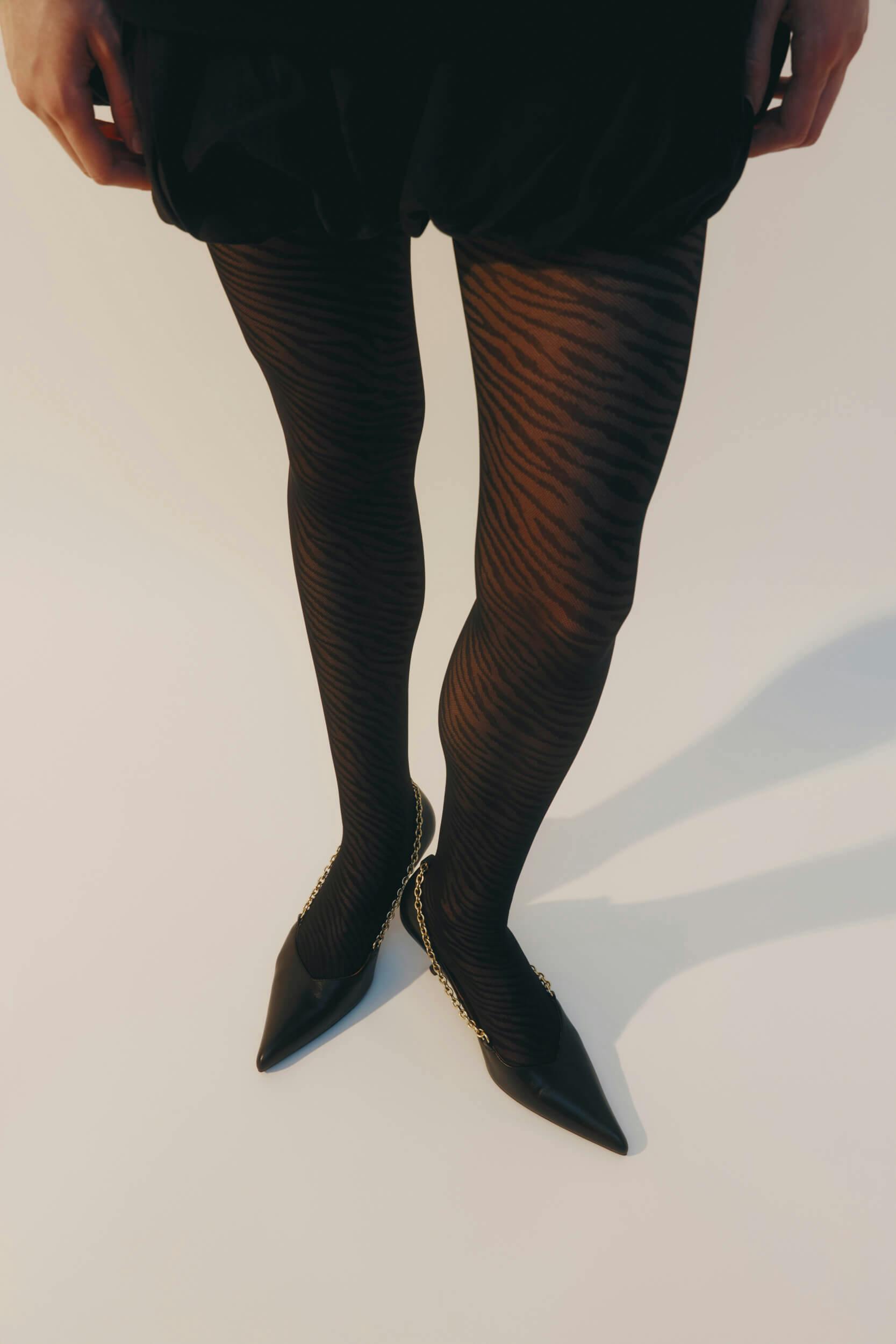 Person wearing black zebra-patterned tights, black shorts, and black pointed-toe heels with gold chain detail, standing against a plain neutral background.