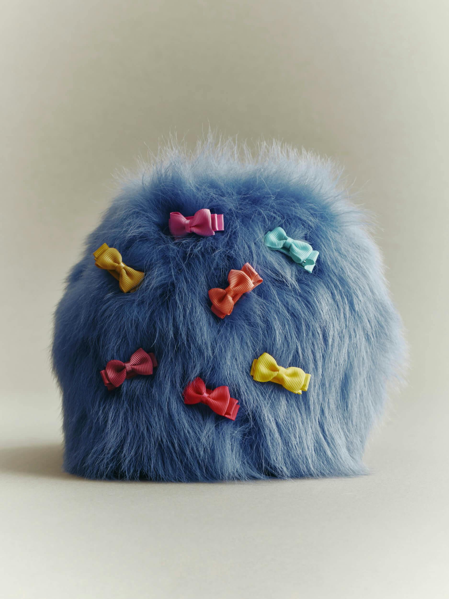 A dome-shaped, fluffy blue object is decorated with seven small, colorful ribbon bows in yellow, pink, red, and light blue, evenly spaced across its front surface. The background is plain and neutral.