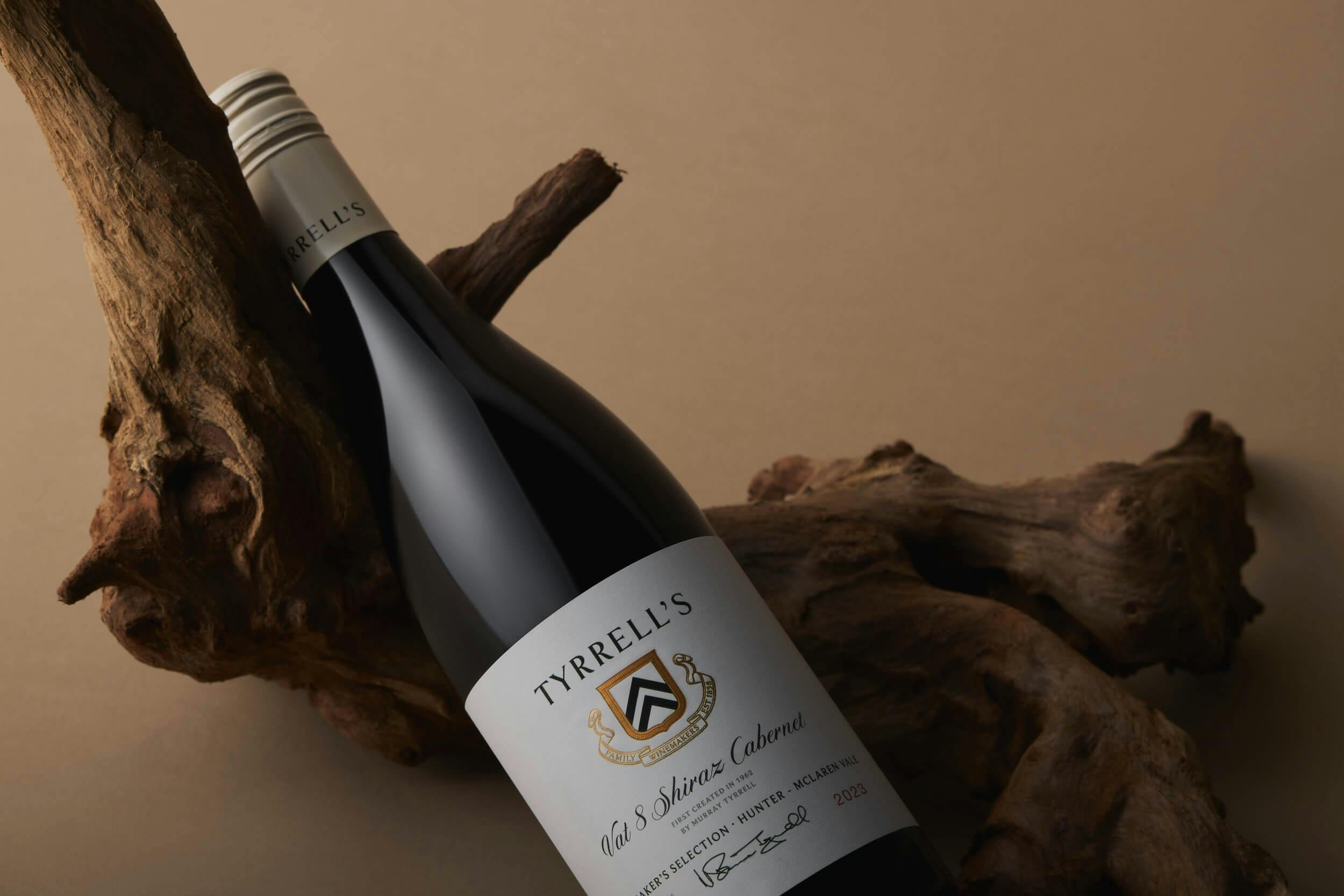 A bottle of Tyrrell's Vat 8 Shiraz Cabernet wine rests on a piece of driftwood against a beige background. The bottle has a white label with text and a gold crest.