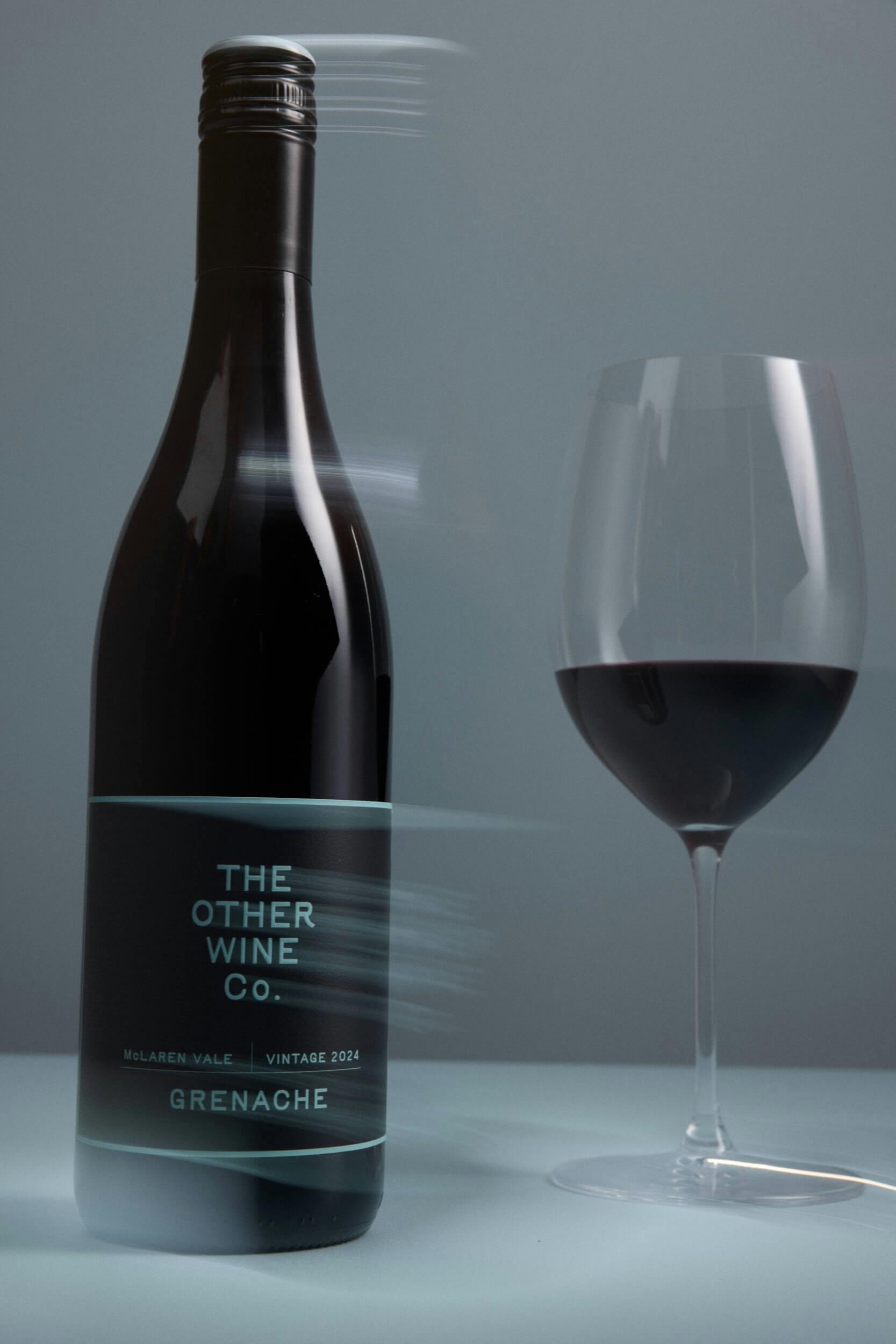 A bottle of The Other Wine Co. Grenache 2024 stands next to a filled glass of red wine on a light blue surface with blurred light streaks in the foreground.