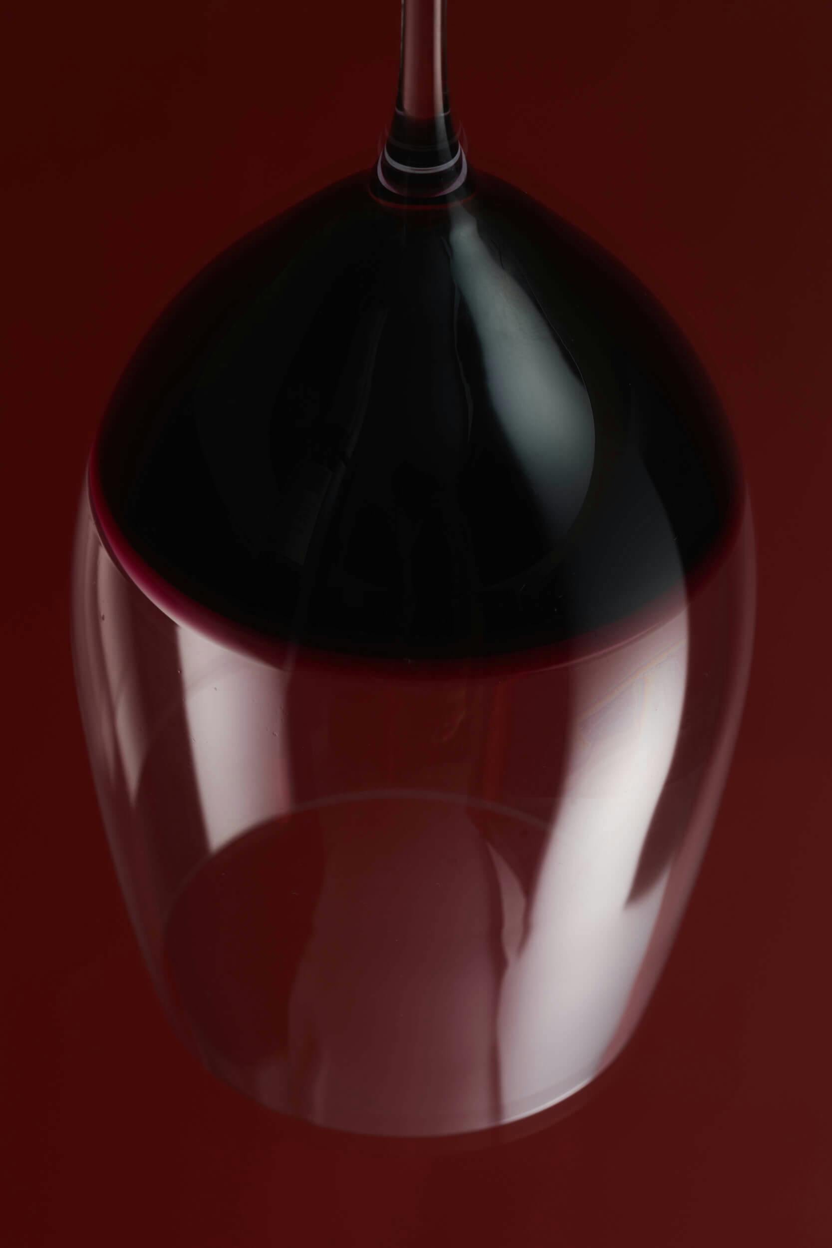 An upside-down wine glass filled with red wine, photographed against a dark red background with reflective highlights on the glass surface.