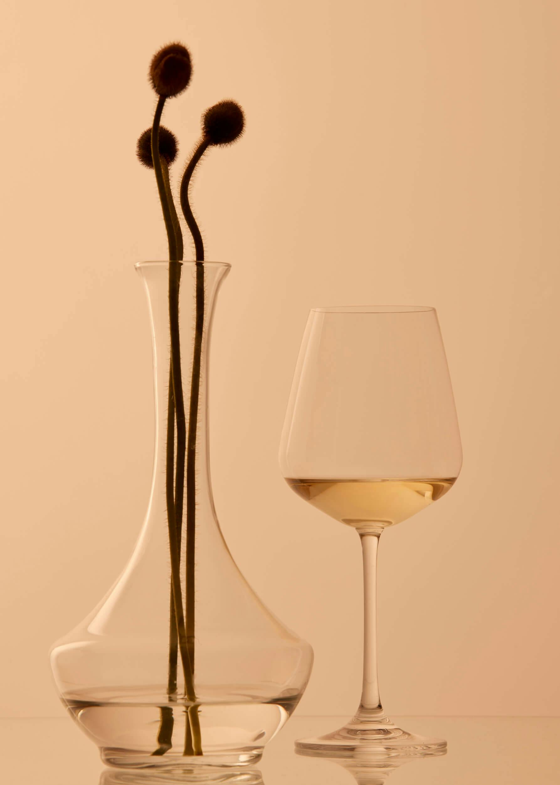 A clear glass vase with three long-stemmed dried flowers next to a wine glass filled with white wine, set against a plain beige background.