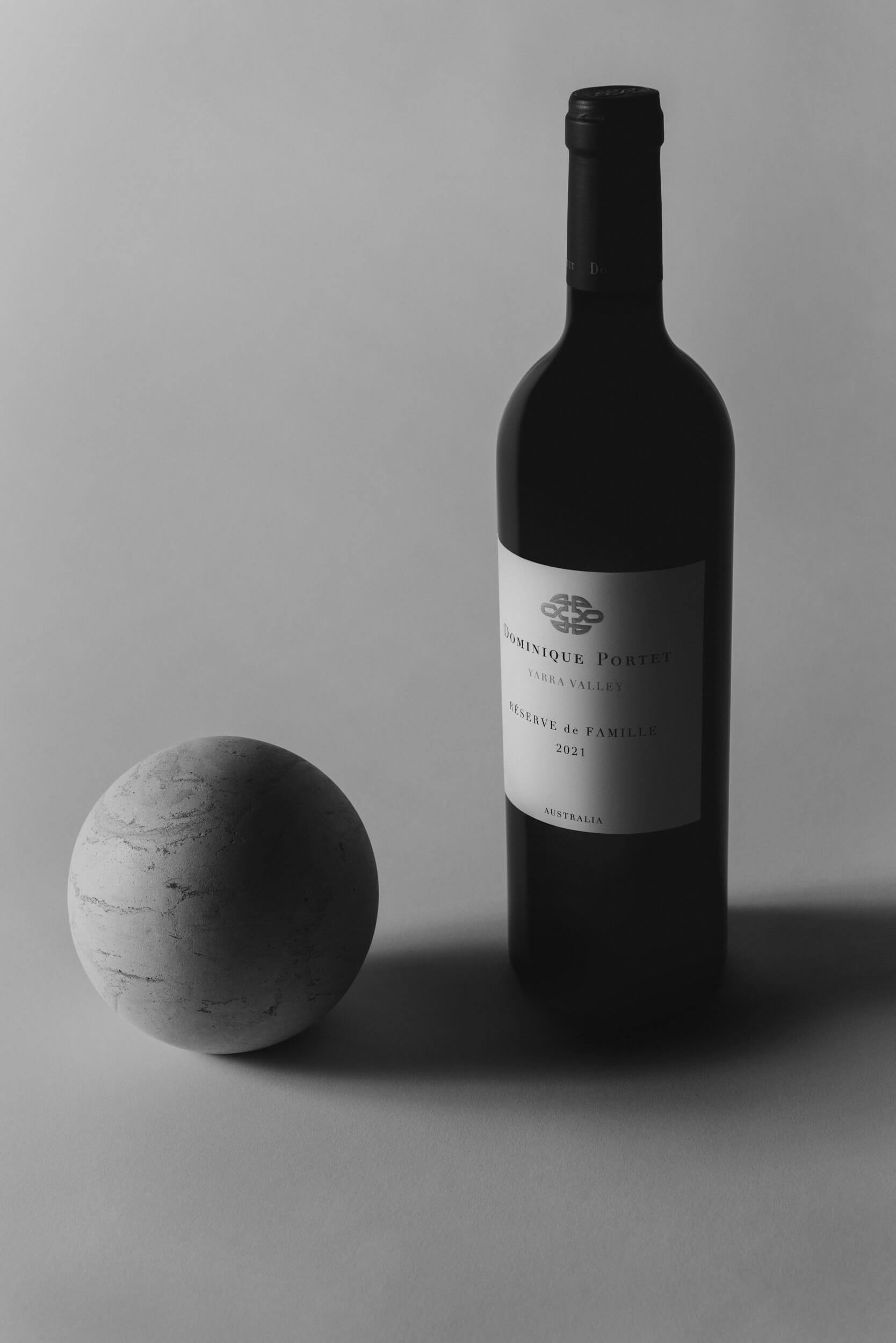 A wine bottle with a label reading “Dominique Portet 2021” stands upright on a plain surface next to a textured stone sphere, both lit by soft, even lighting in a black-and-white photo.