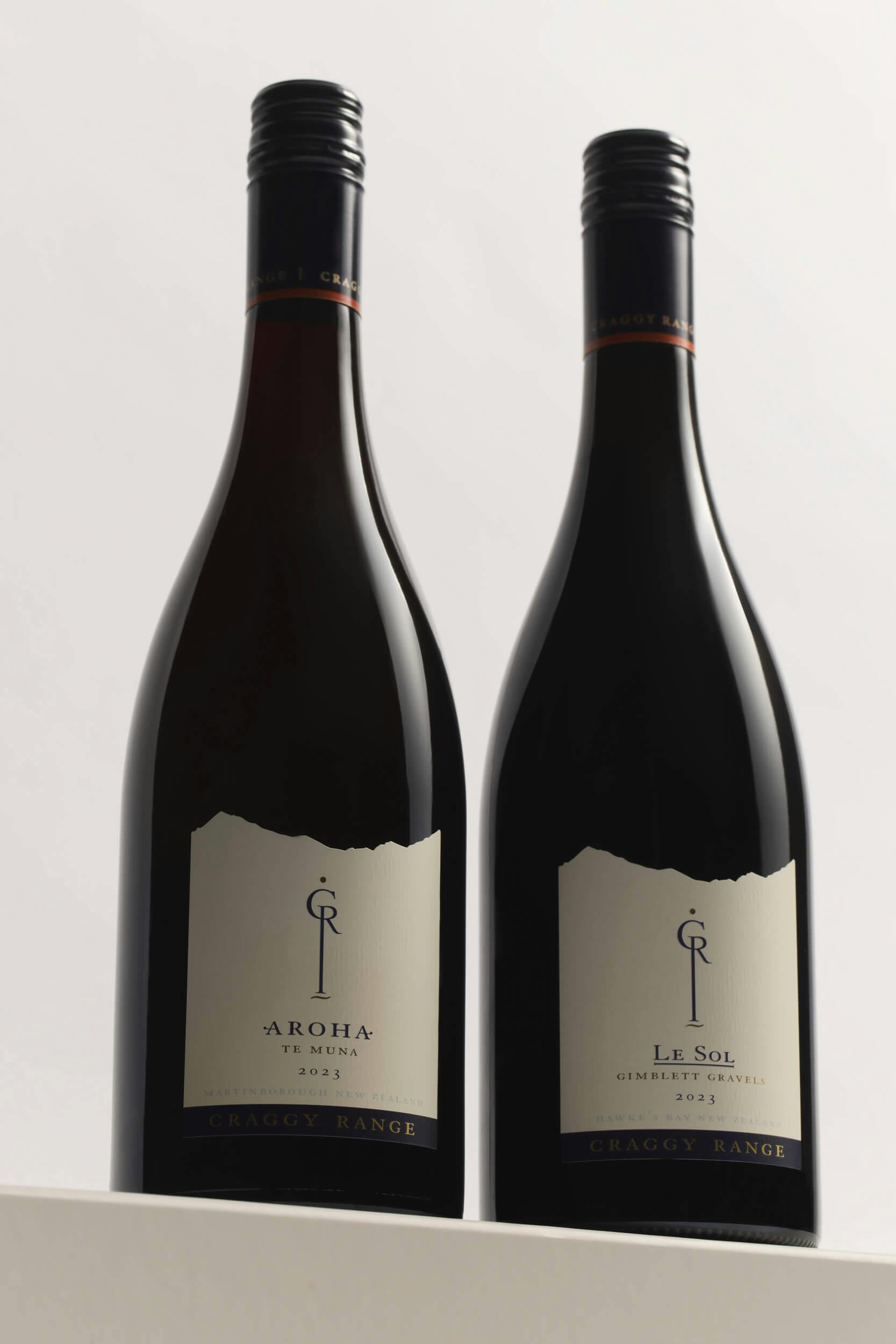 Two bottles of red wine stand side by side on a white surface. The left bottle is labeled 