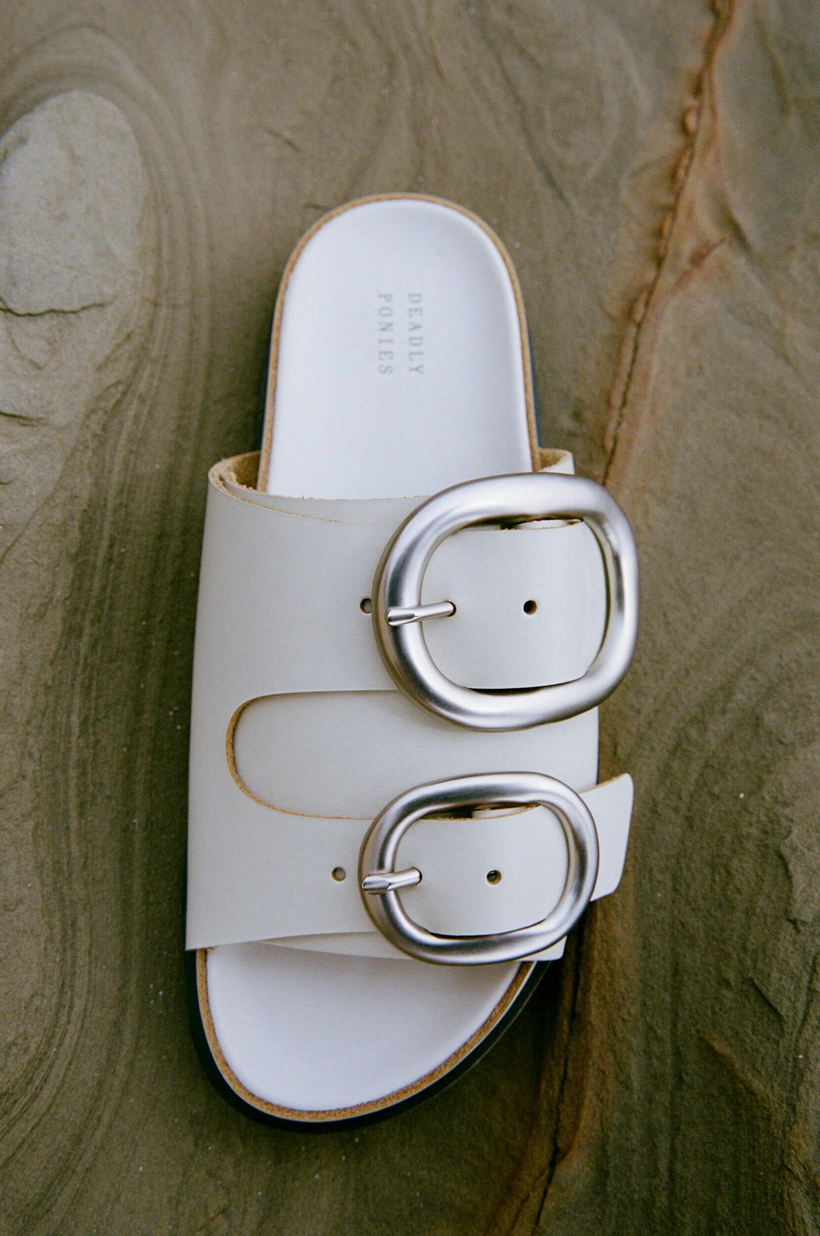 A white slide sandal with two large silver buckles is placed on a textured stone surface. The sandal has the words