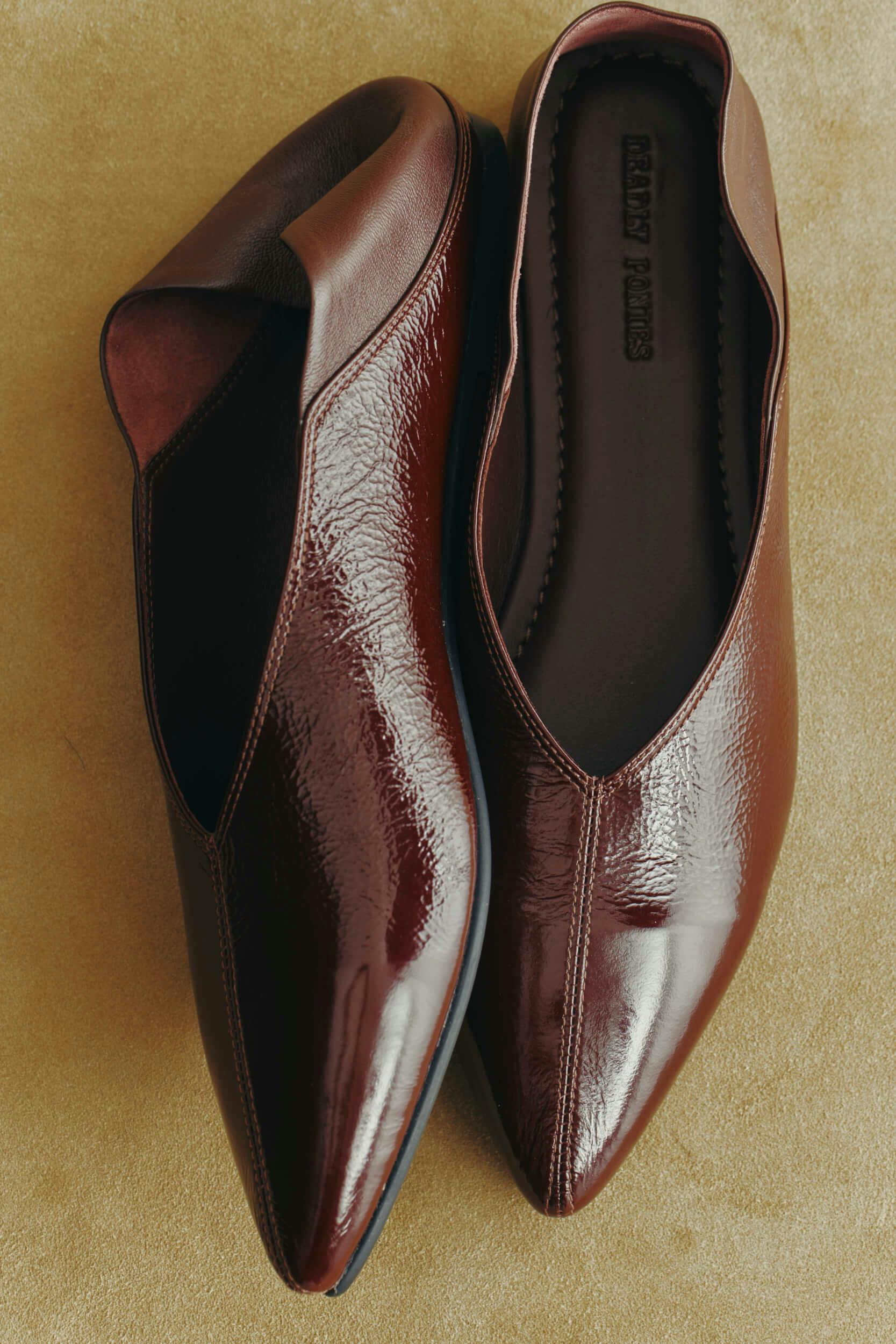 A pair of pointed-toe brown leather flats is placed side by side on a beige surface, with subtle texture and shine visible on the leather.