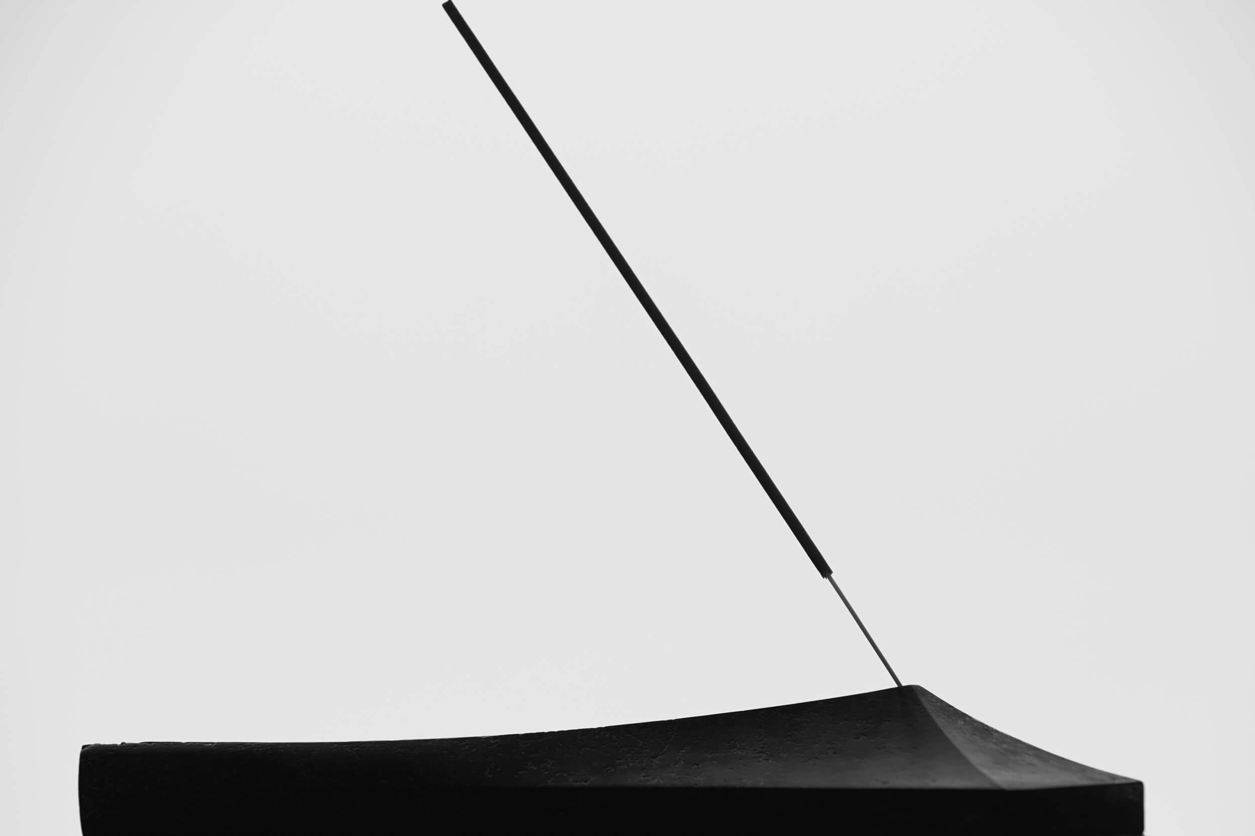 A stick of incense is positioned at an angle on a curved black surface, with smoke barely visible against a plain white background. The image is minimalistic with high contrast and simple shapes.
