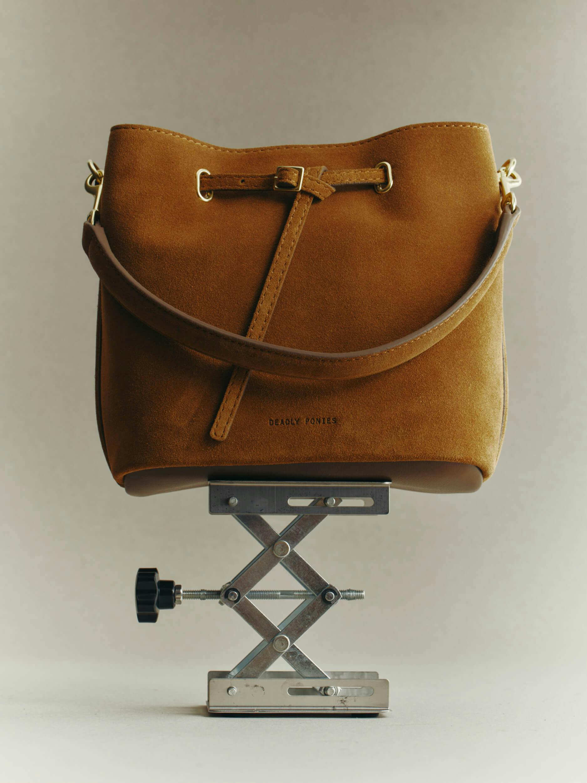 A brown suede handbag with a shoulder strap is displayed upright on a metal scissor lift against a plain, light background.