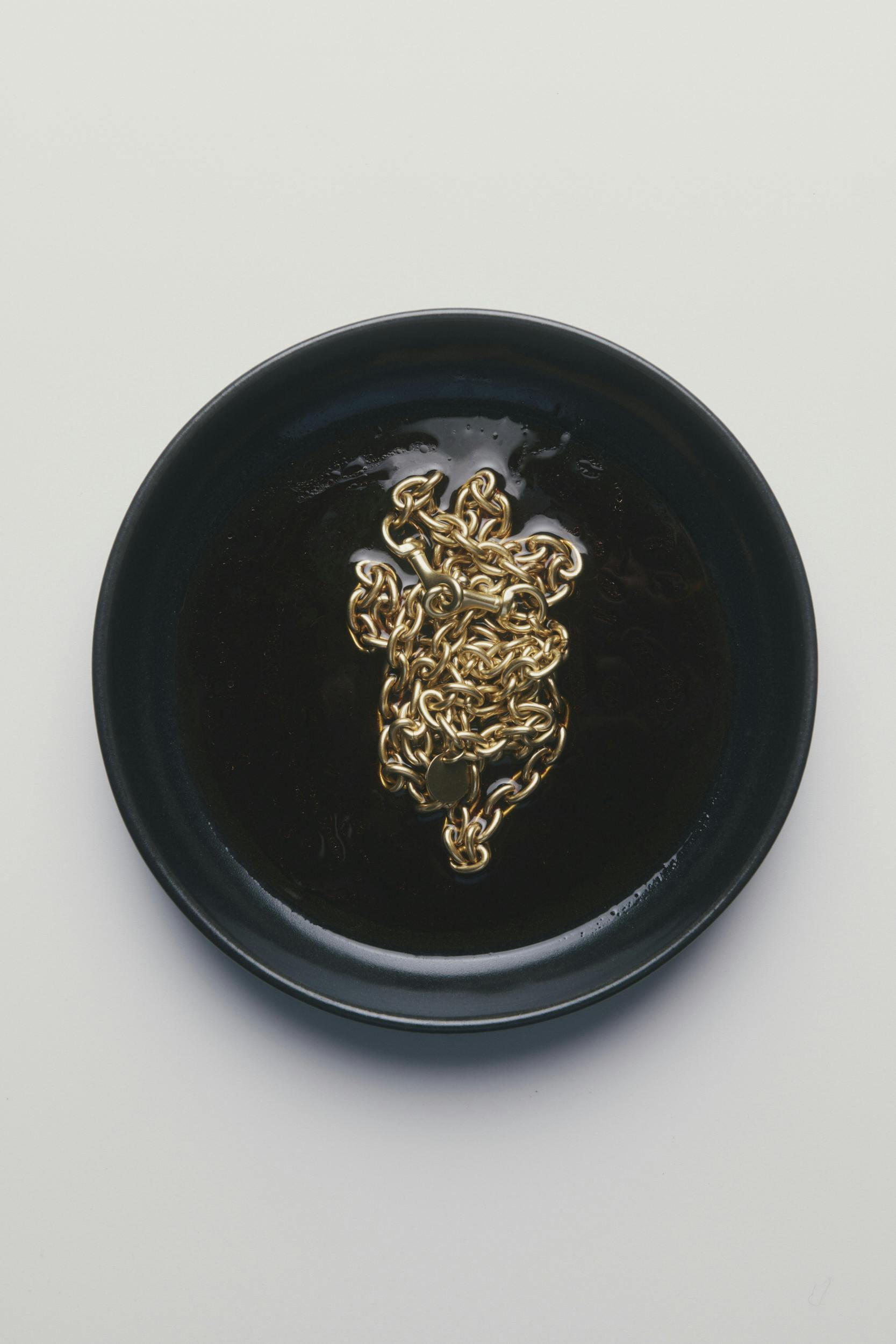 A gold chain is coiled in the center of a black plate, which is placed on a plain white background.