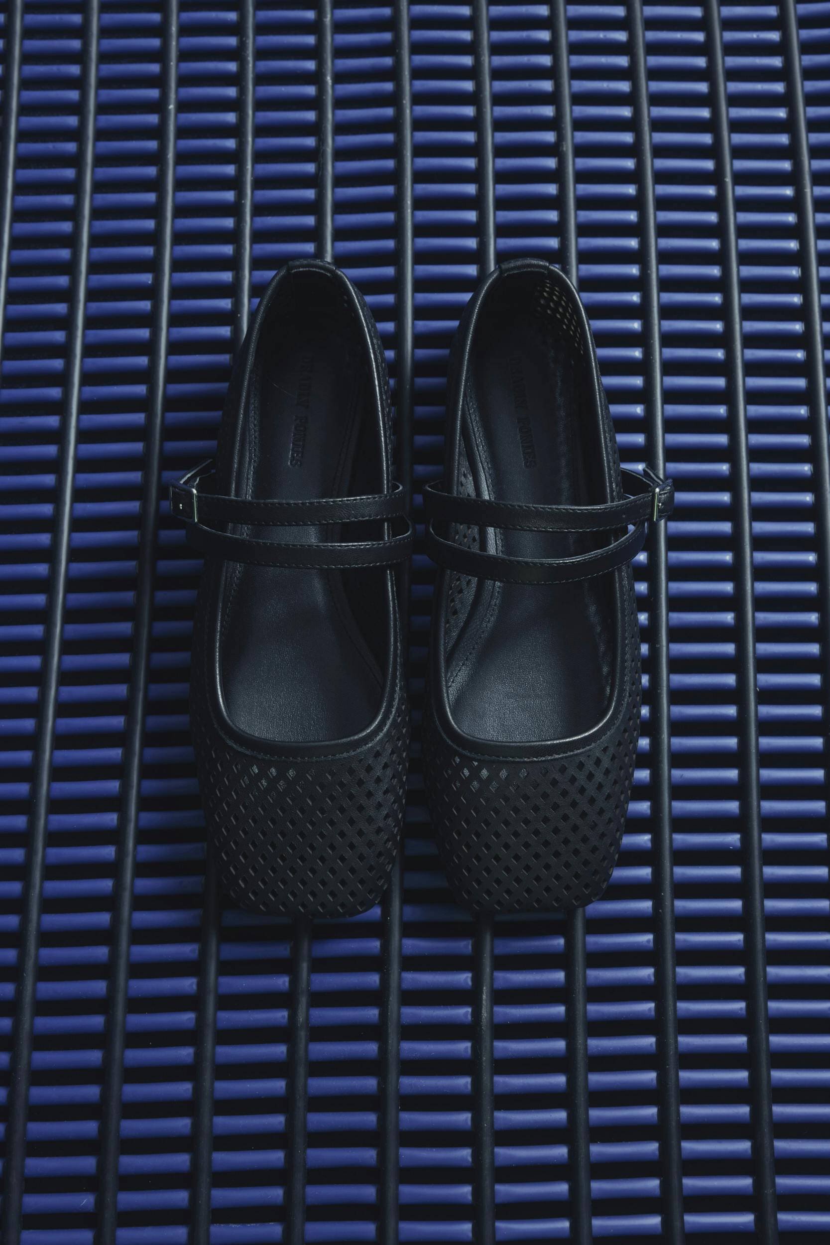 A pair of black, perforated, square-toed Mary Jane shoes with straps are placed side by side on a blue slatted surface.