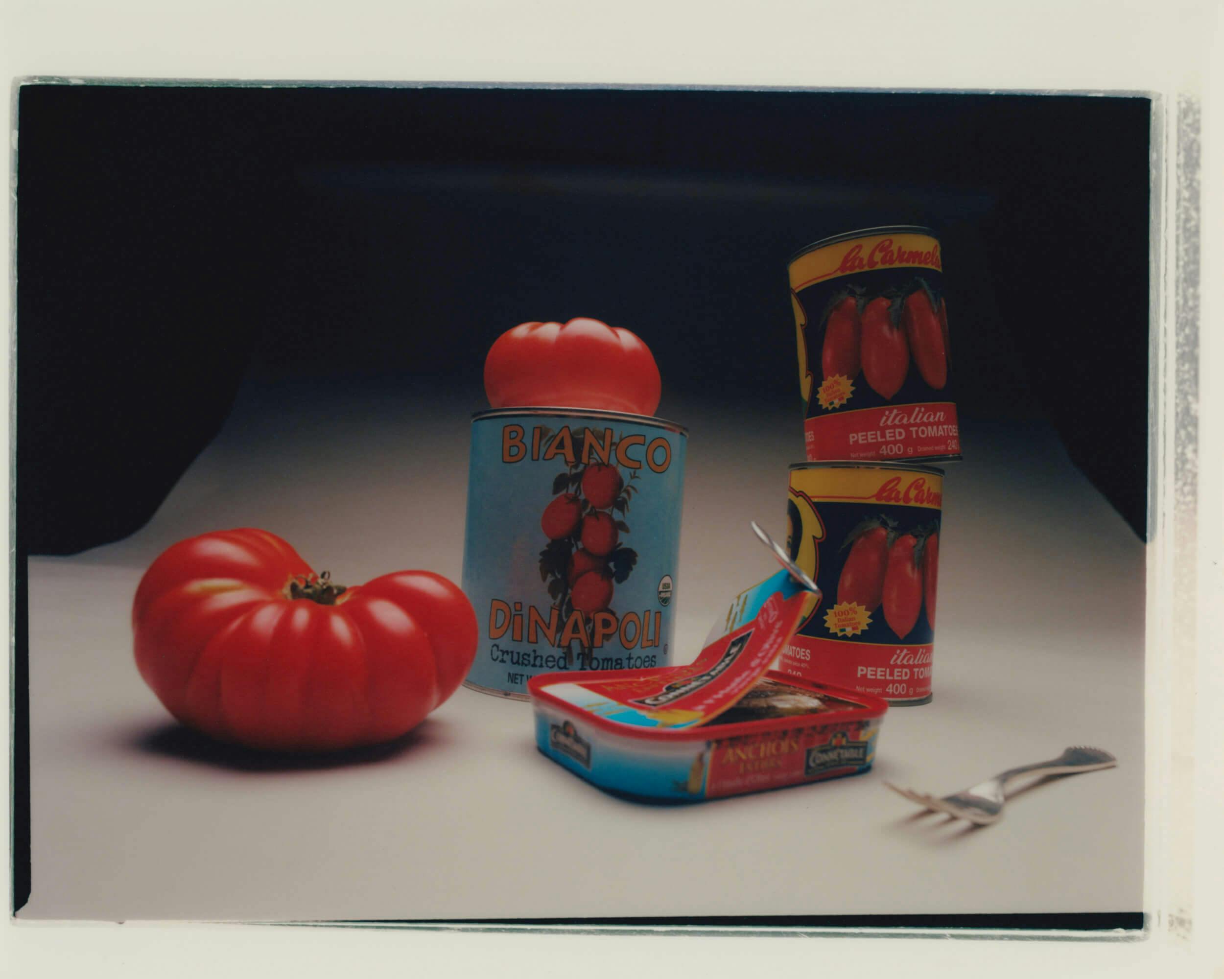 A tomato sits next to an opened tin of anchovies, a fork, and three cans of peeled or crushed tomatoes, arranged on a white and black surface.