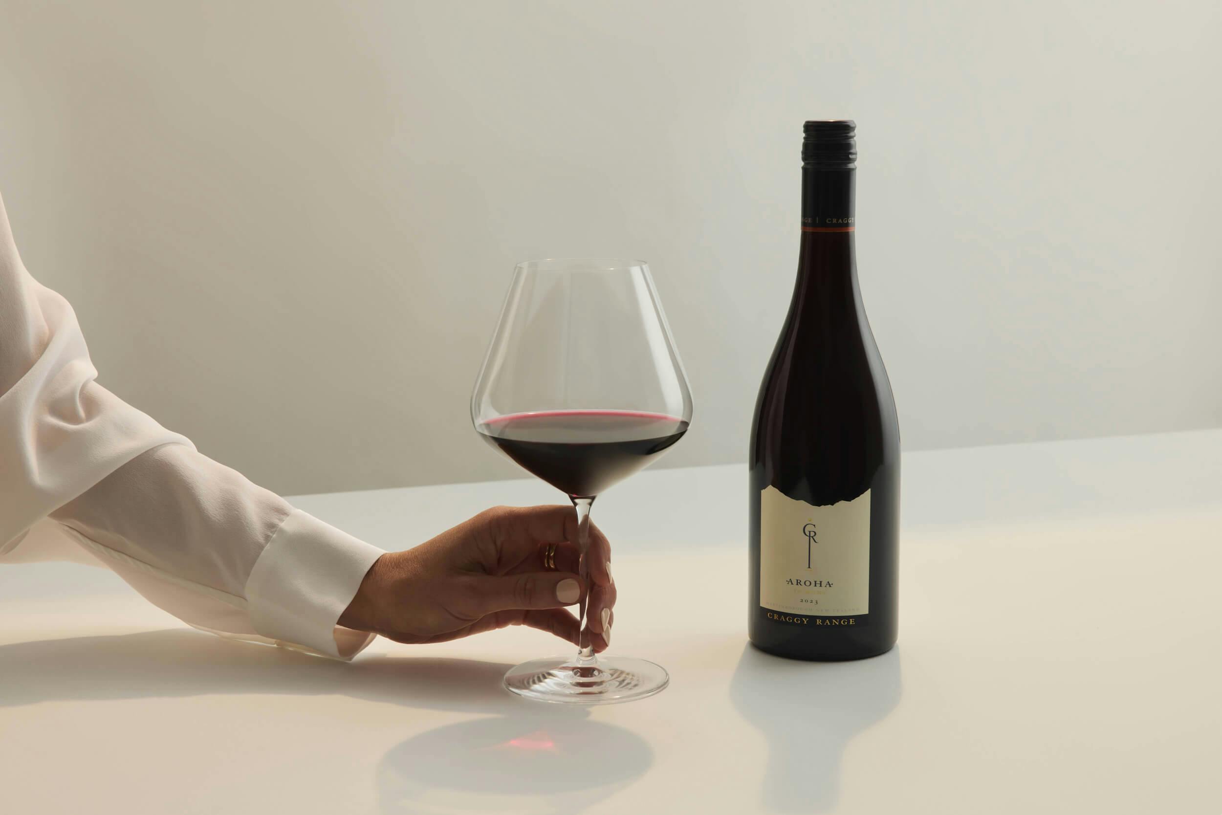A person wearing a white long-sleeve shirt holds a glass of red wine next to a bottle of wine with a dark label, both placed on a white surface against a plain light background.