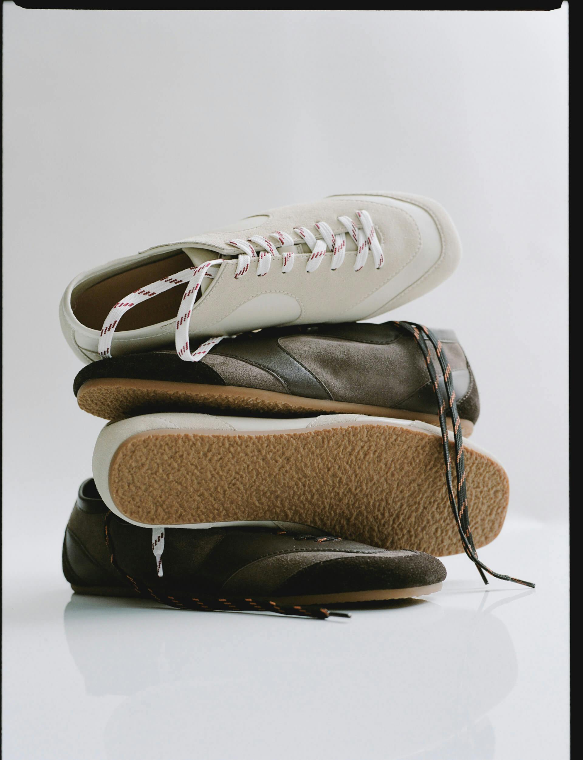Three sneakers are stacked on top of one another against a plain white background. The top sneaker is white with light brown soles, and the two lower sneakers are dark brown with gum soles. Shoelaces are visible on all pairs.