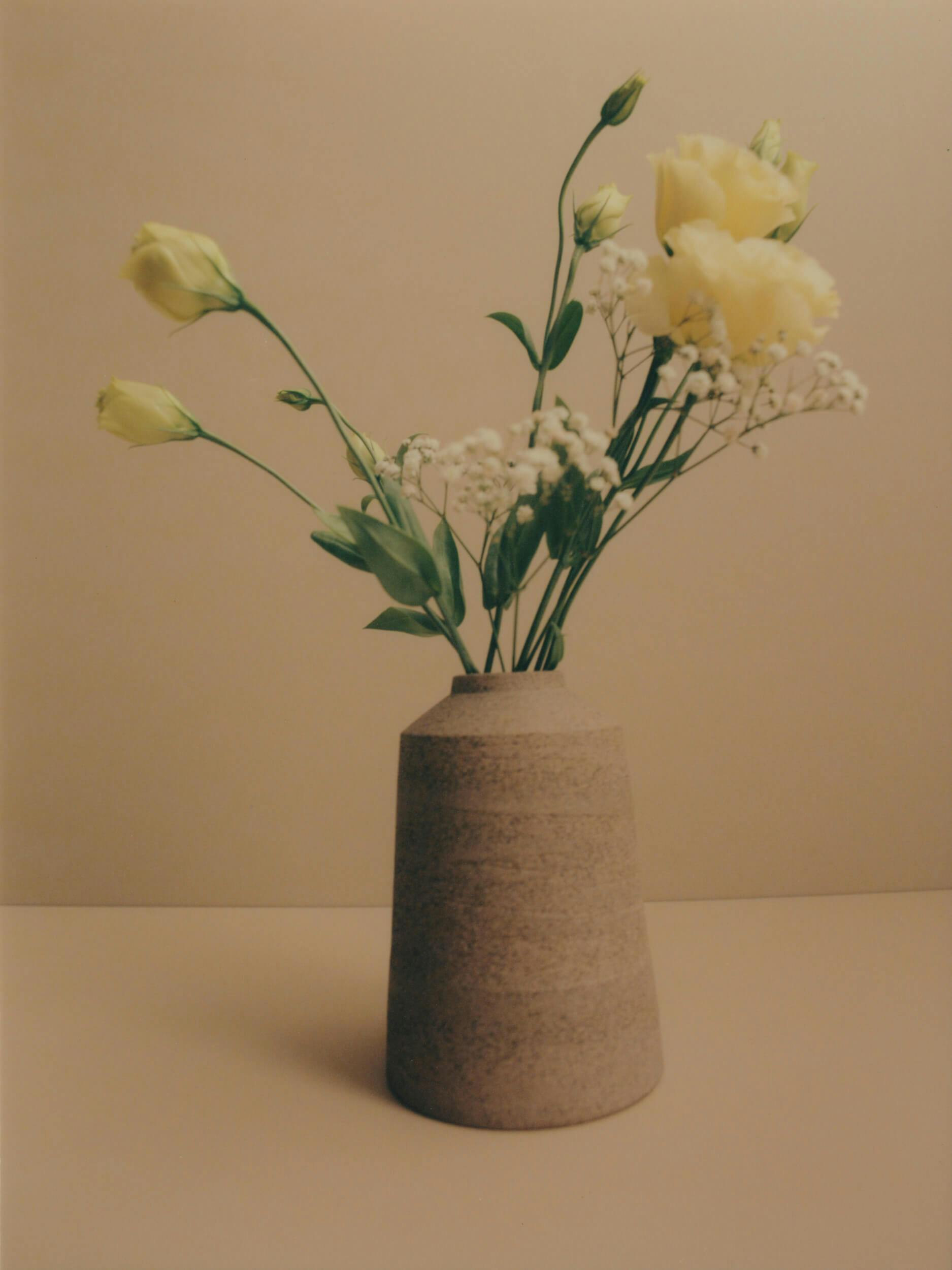 A beige ceramic vase holds light yellow flowers, small white flowers, and green leaves, set against a neutral beige background.