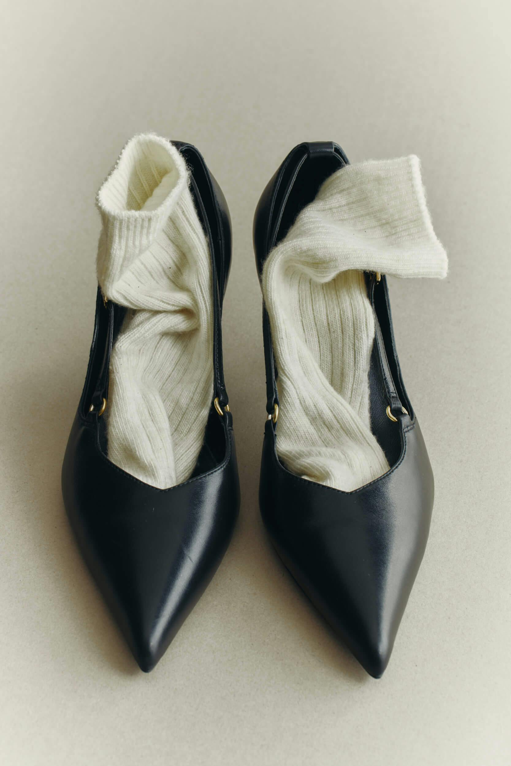 A pair of black pointed-toe high-heeled shoes with white ribbed socks loosely placed inside each shoe, set on a light grey surface.