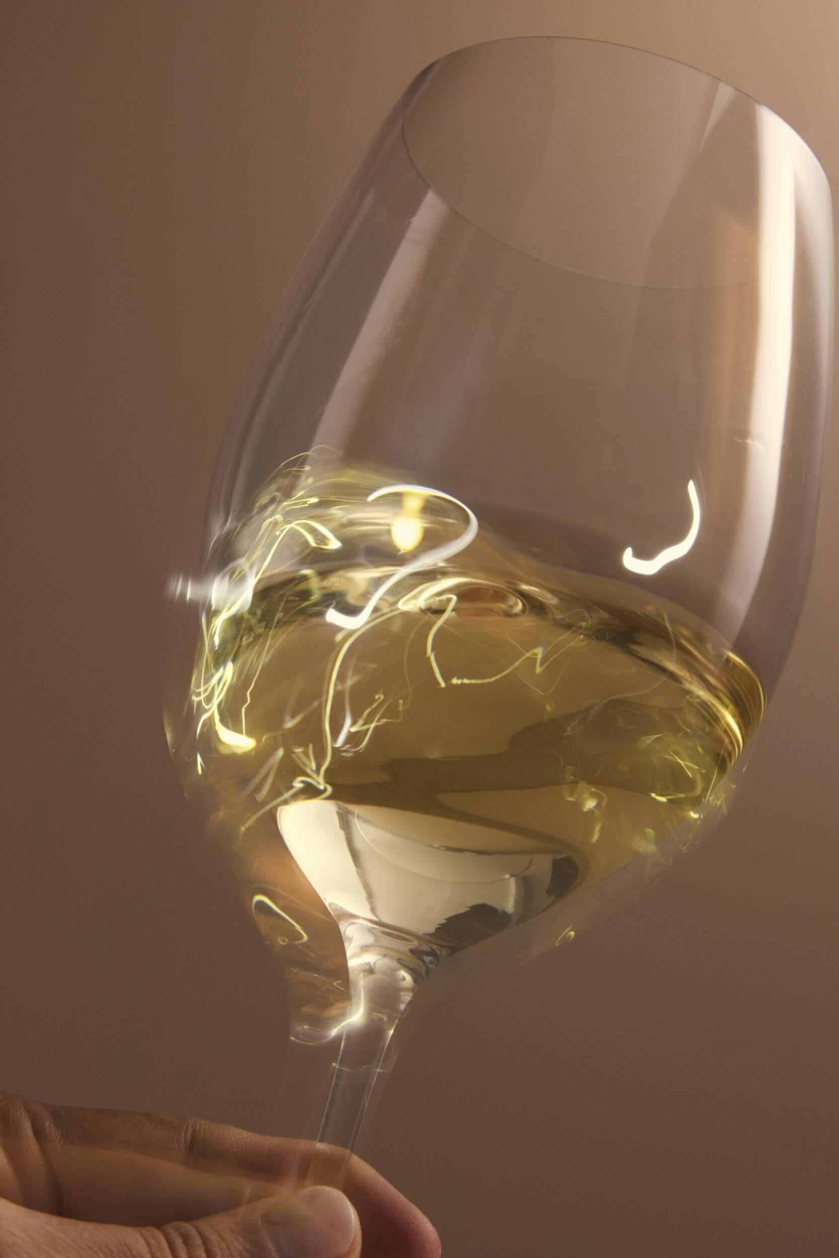 A close-up of a hand holding a tilted wine glass filled with white wine, showing reflections and highlights on the glass surface against a neutral, softly lit background.