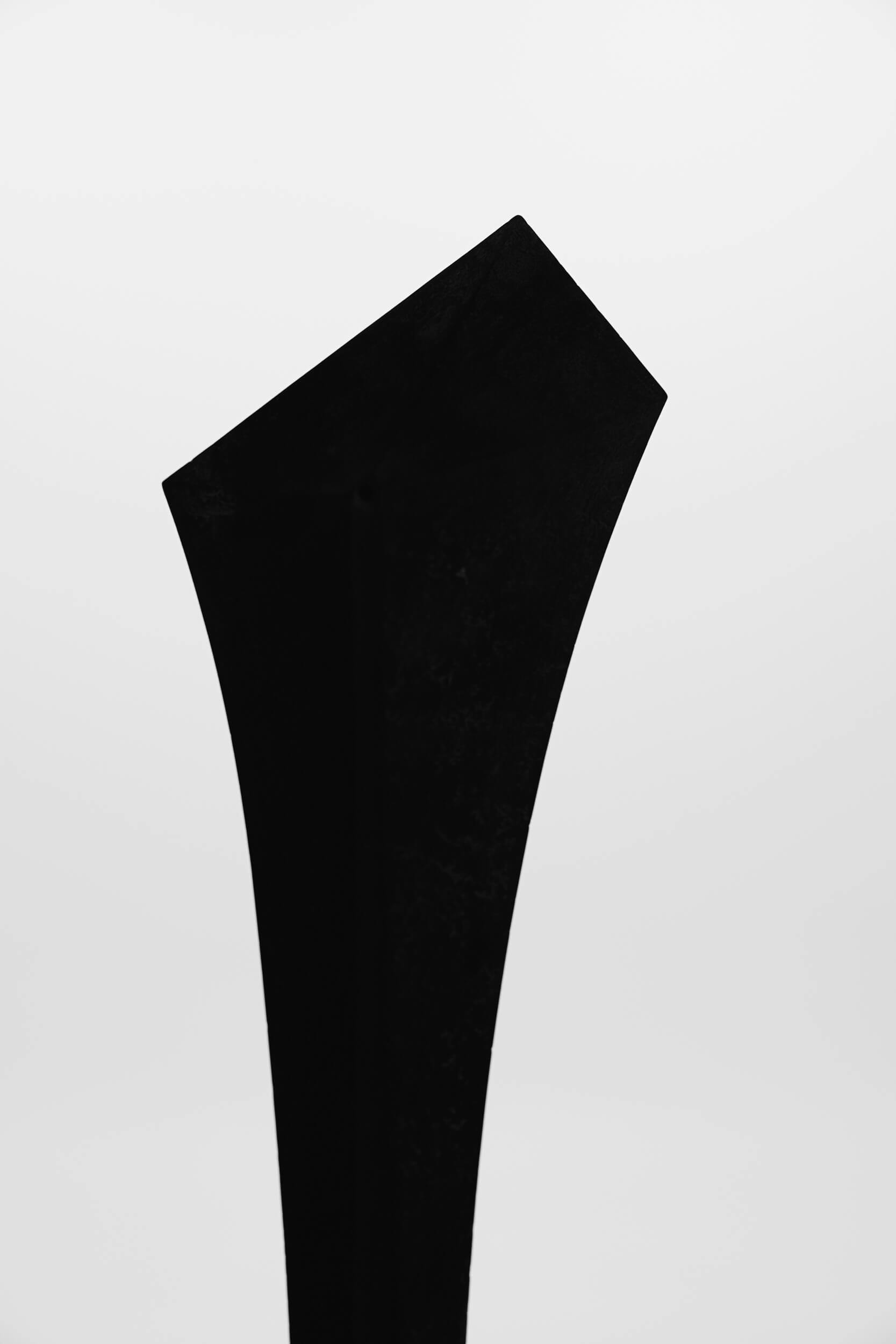 A tall, black, angular abstract sculpture with a geometric, pointed top standing against a plain white background.
