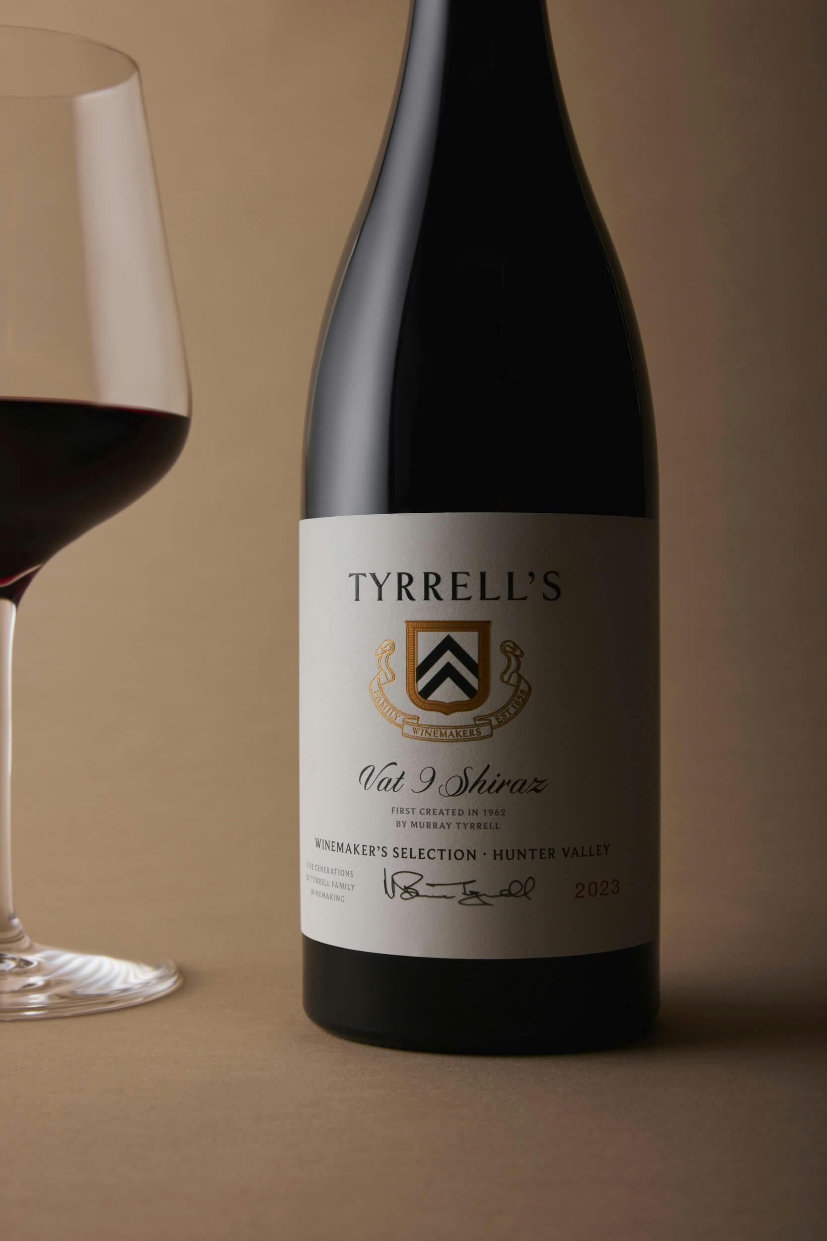 A bottle of Tyrrell's Val Shiraz wine stands next to a partially filled wine glass, both set against a neutral beige background. The wine label displays the vintage year 2023 and details about the winery.